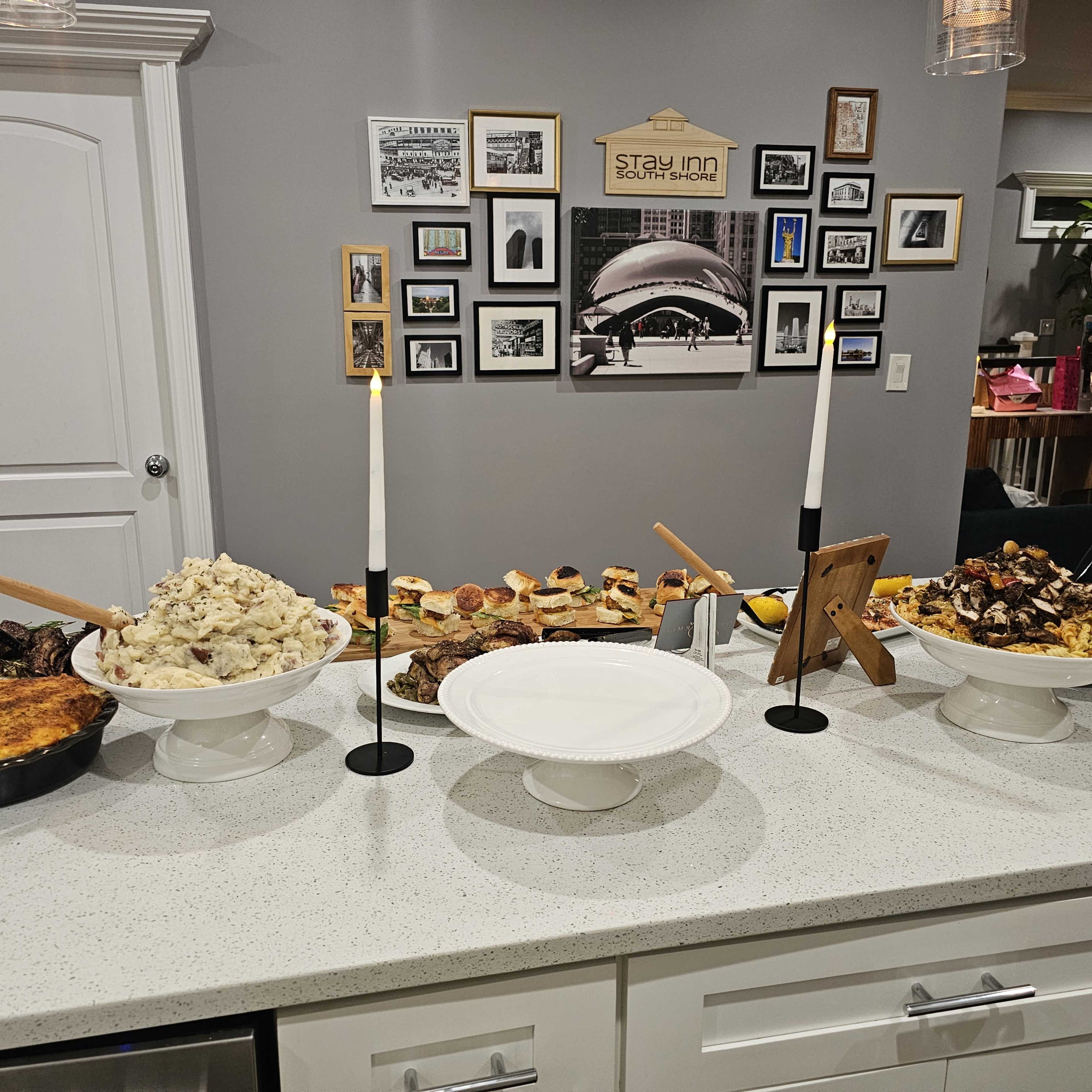 A buffet spread on a white countertop featuring various dishes, including meats, mashed potatoes, appetizers, and candle holders, with a wall of framed photos in the background.