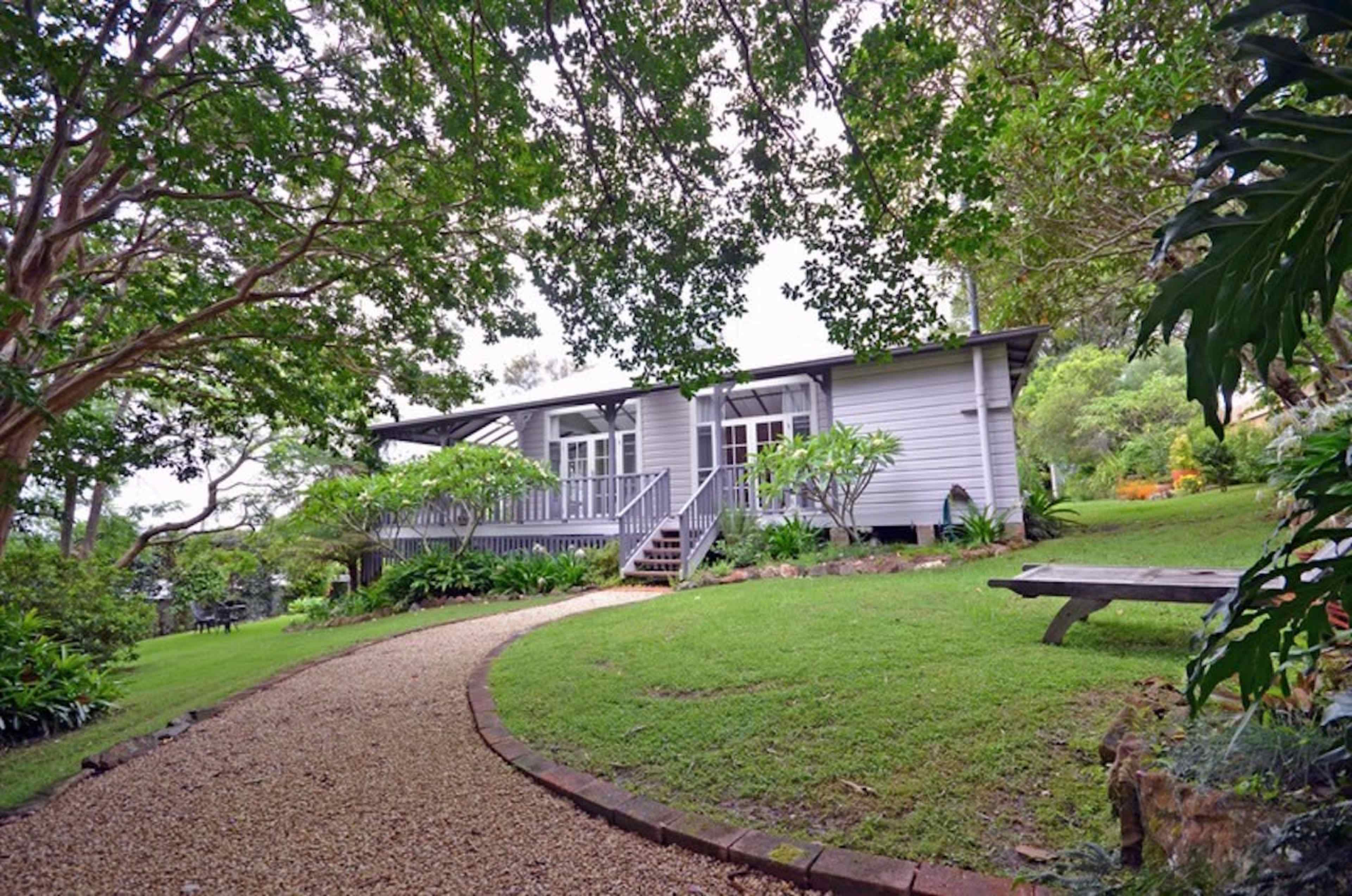 A single-story house with large windows is surrounded by lush greenery and a gravel pathway leading to the entrance.