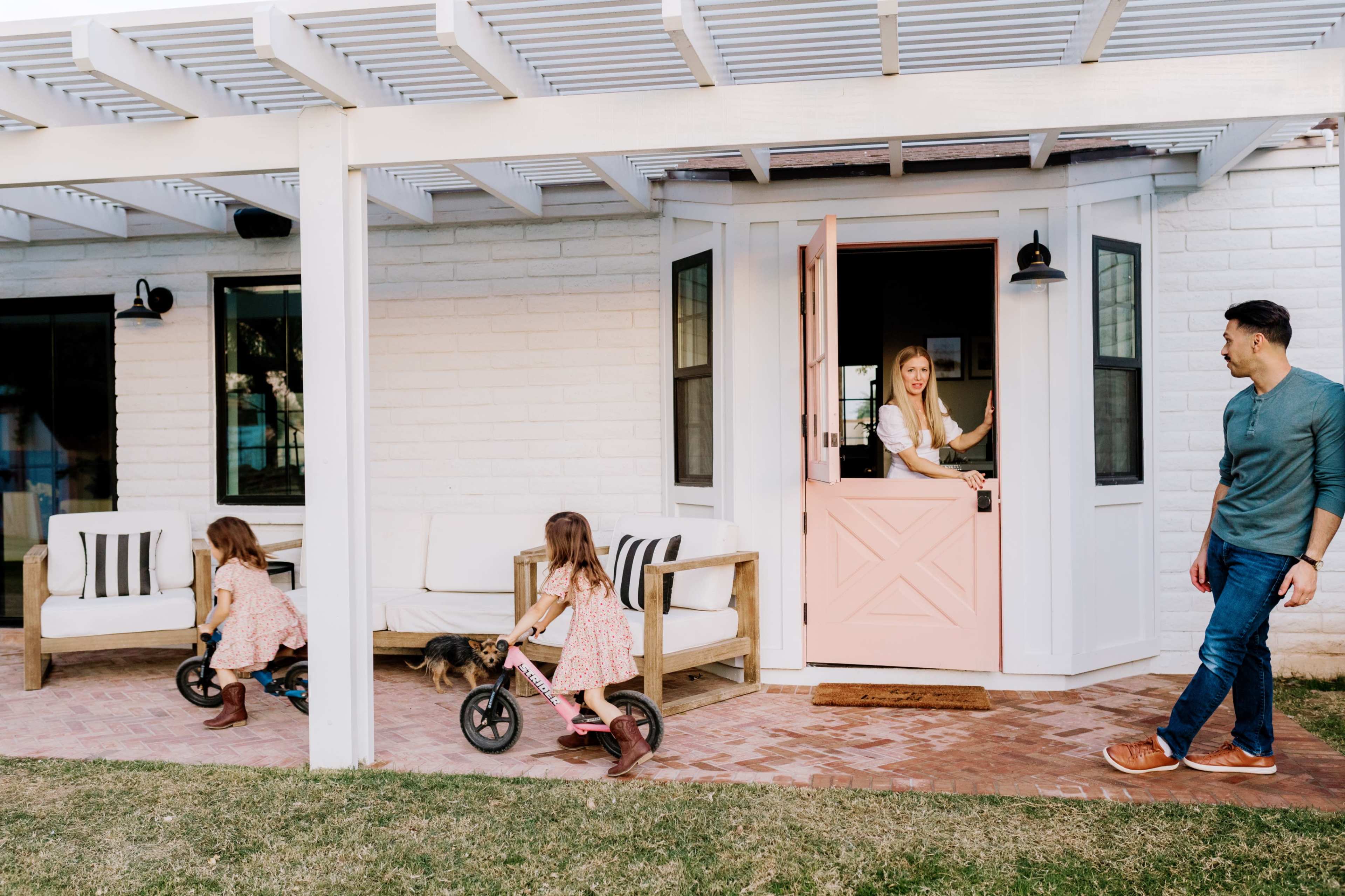 A woman stands in a pink doorframe while two young girls ride small bikes on a brick patio outside a white house.