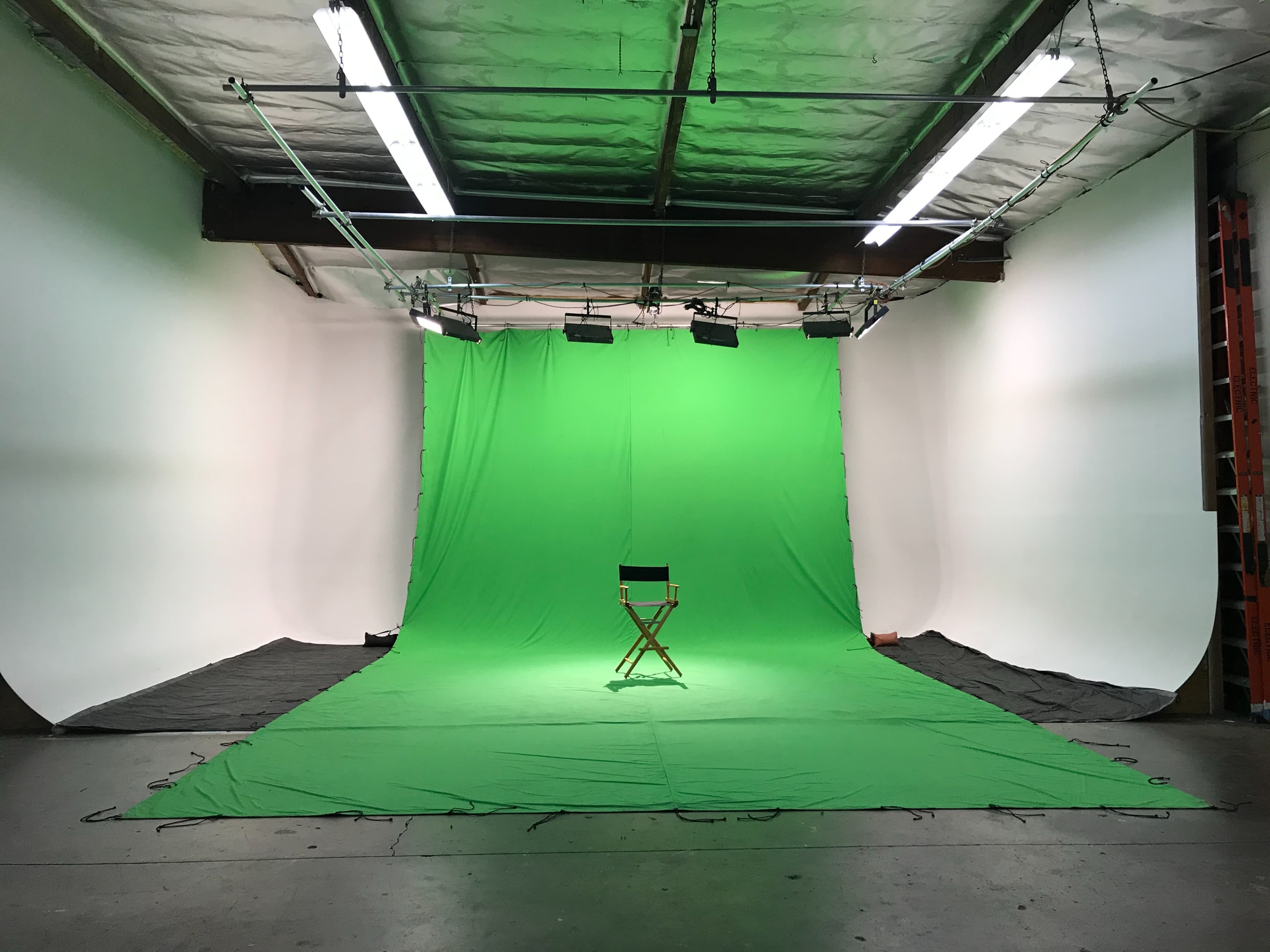 A green screen studio features a central director's chair on a flat surface surrounded by green fabric and white backdrops.