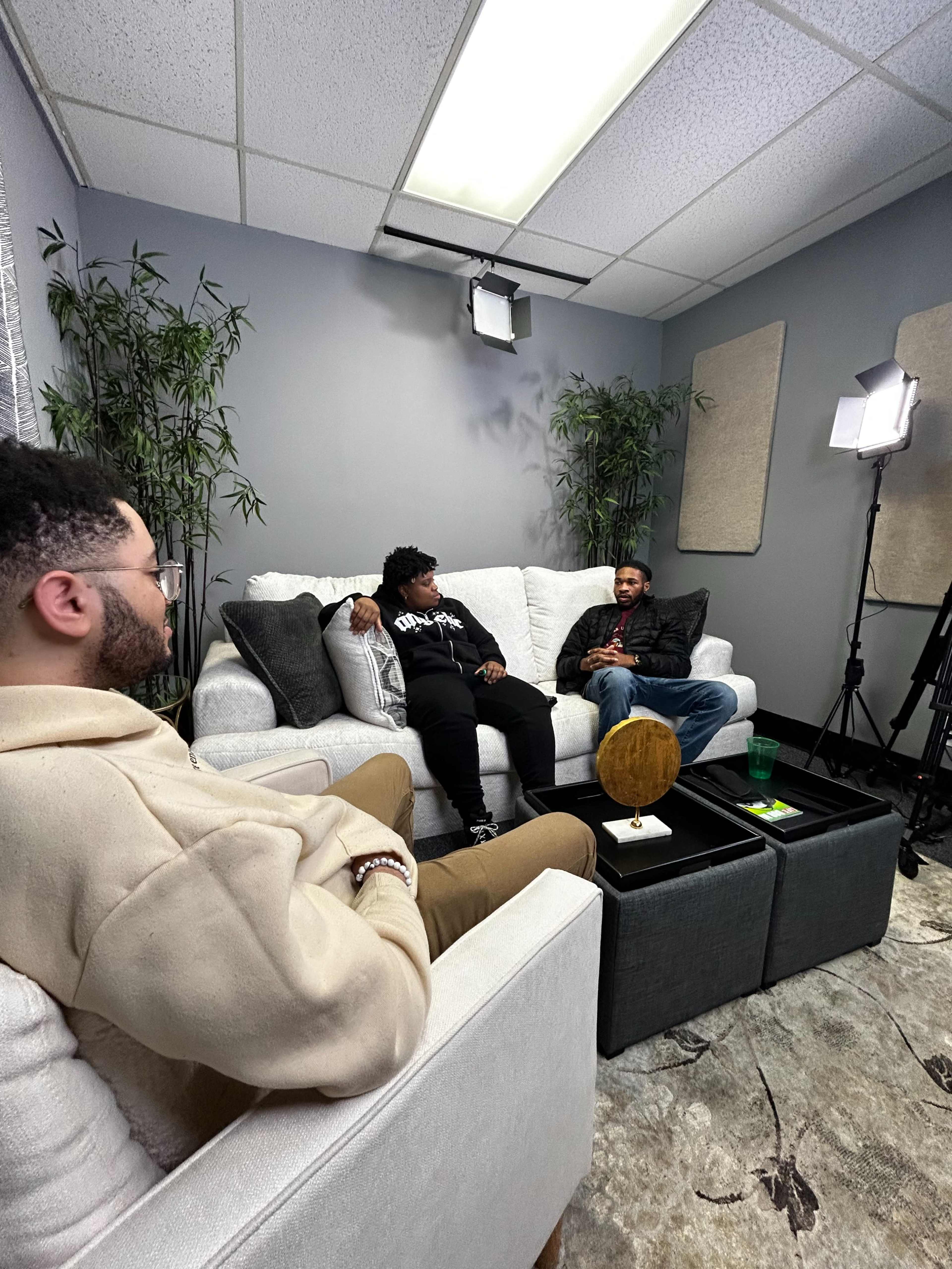 Three individuals are seated on a white couch in a well-lit room with greenery, discussing amidst studio lighting equipment.