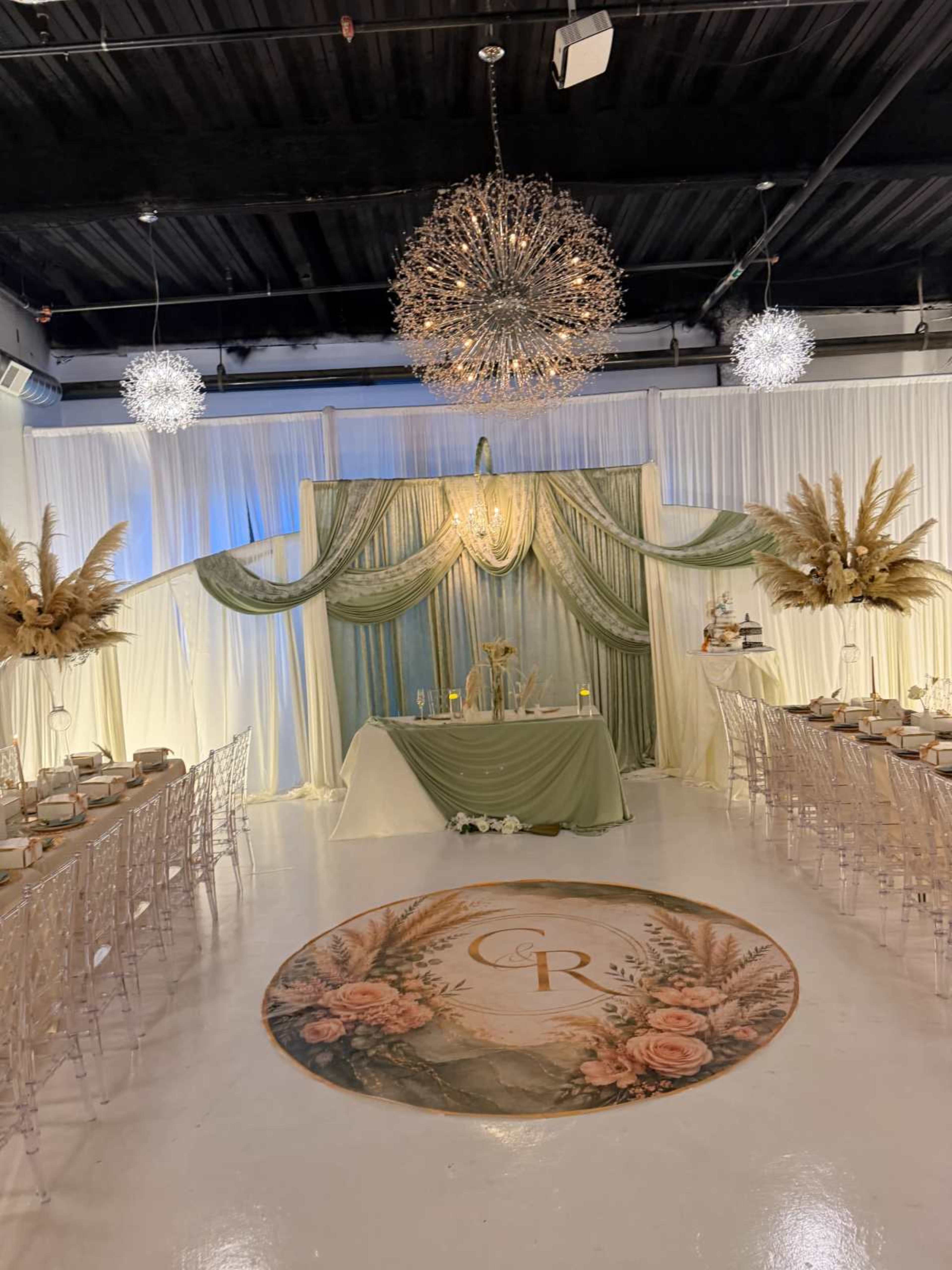 The image shows a decorated event space with an elaborate backdrop featuring draped fabric and floral arrangements, flanked by elegantly set tables and chandeliers overhead.