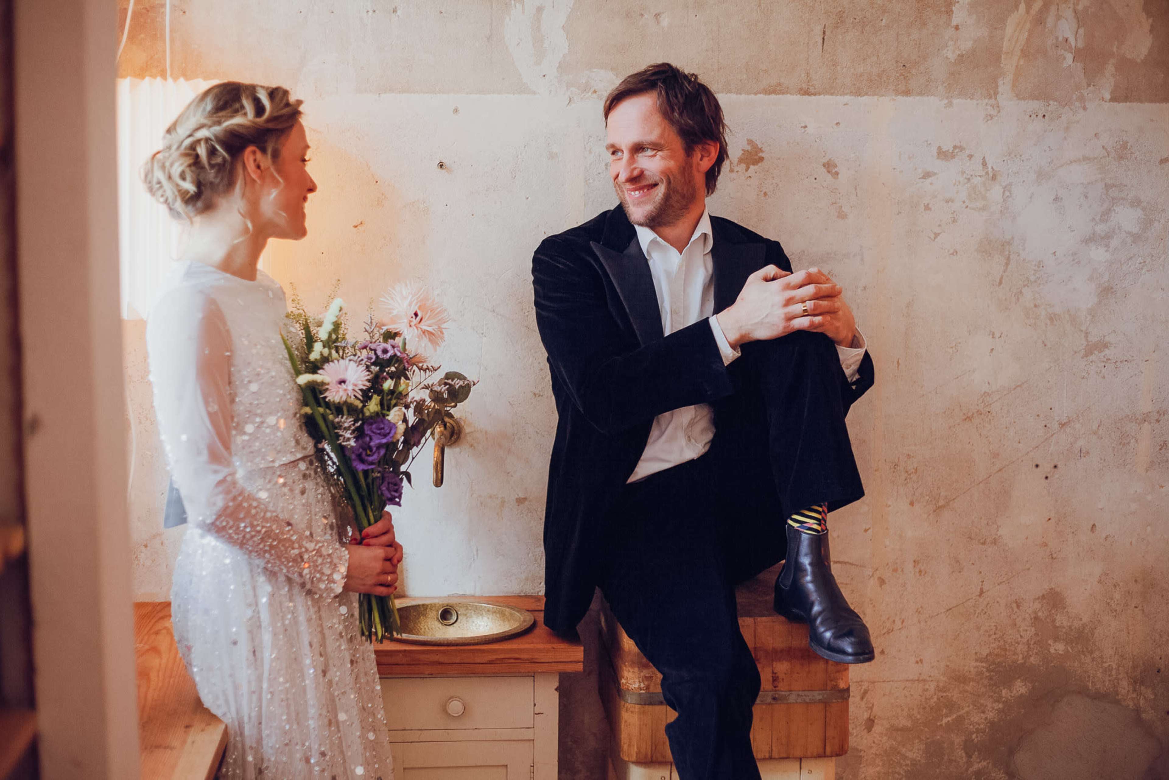 A woman in a sequined dress stands holding a bouquet of flowers while a man in a black jacket sits on a wooden crate, both smiling at each other in a rustic room.