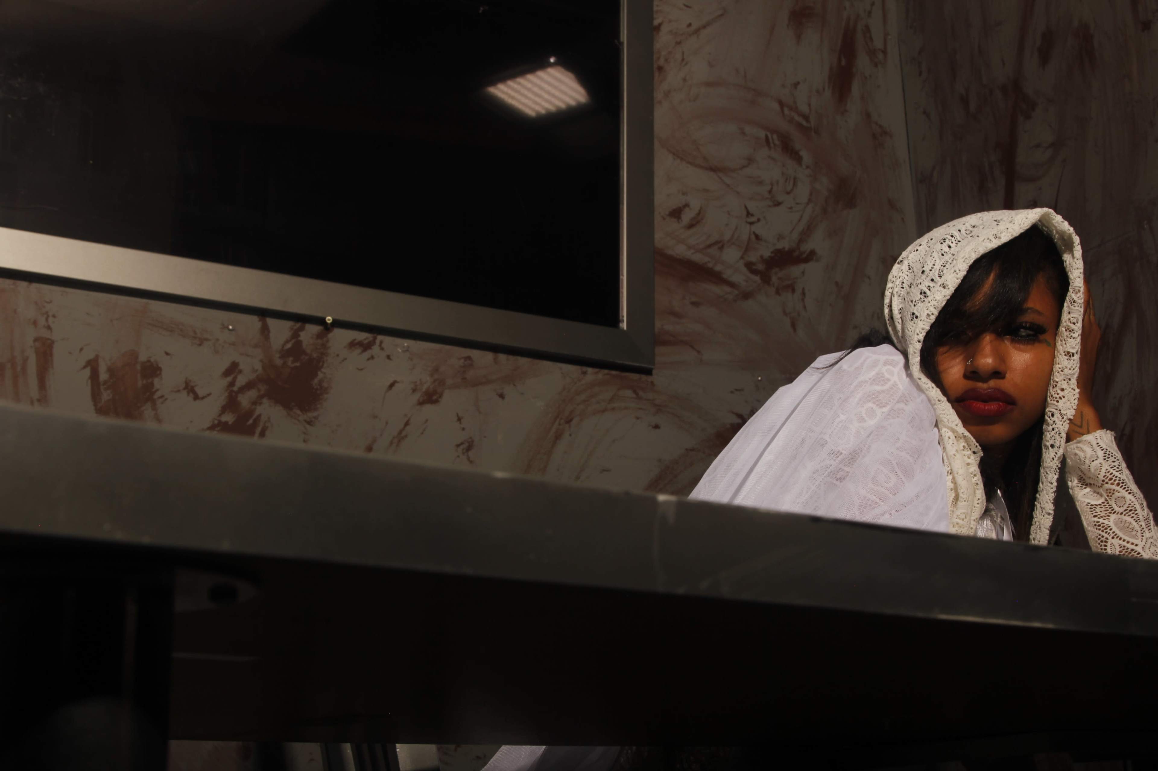 A woman with a headscarf sits at a dark table, leaning against her hand while looking towards a monitor in a room with textured walls.