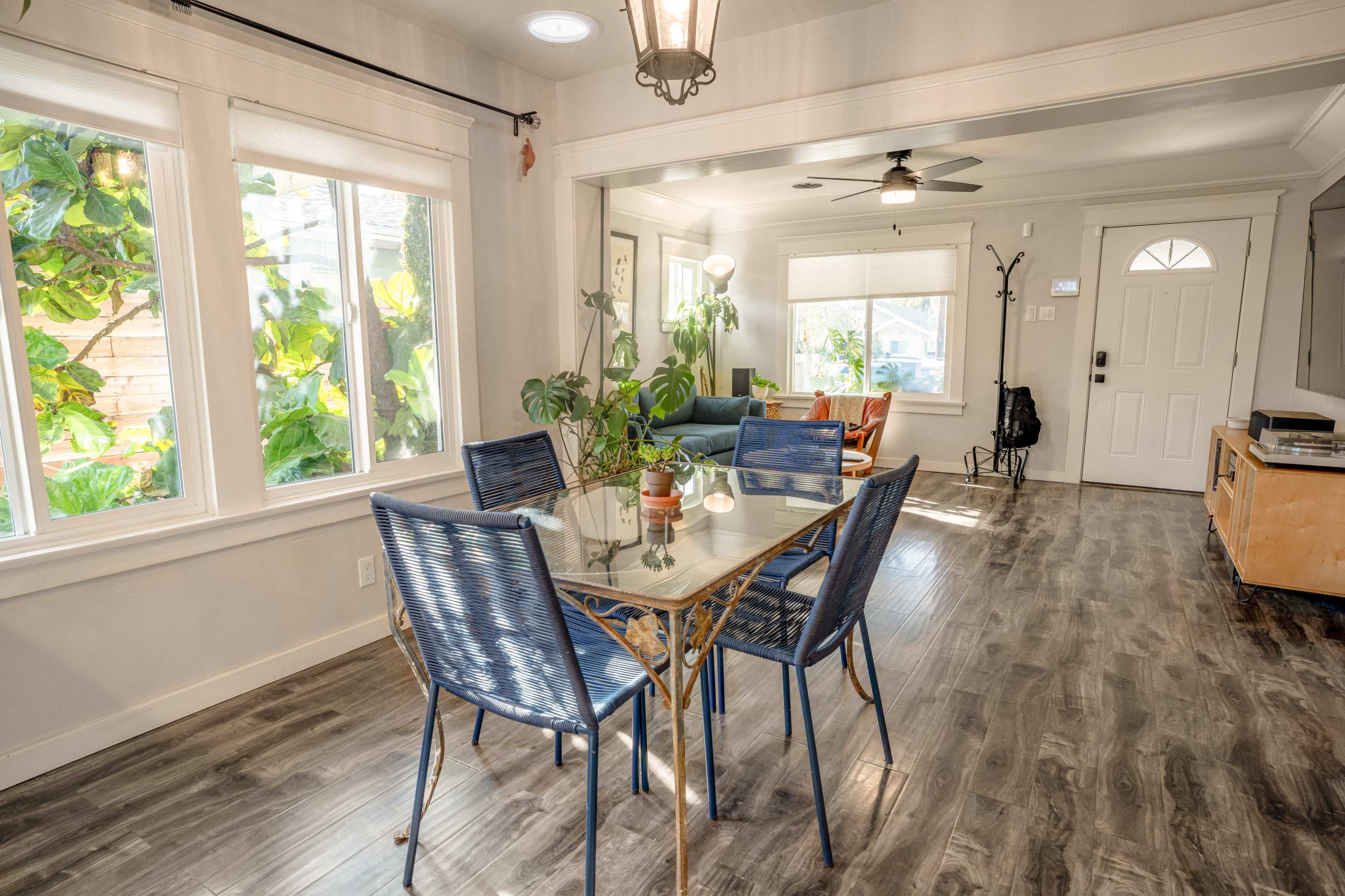 A dining table with blue chairs is positioned near large windows, overlooking a garden, with a living area visible in the background.