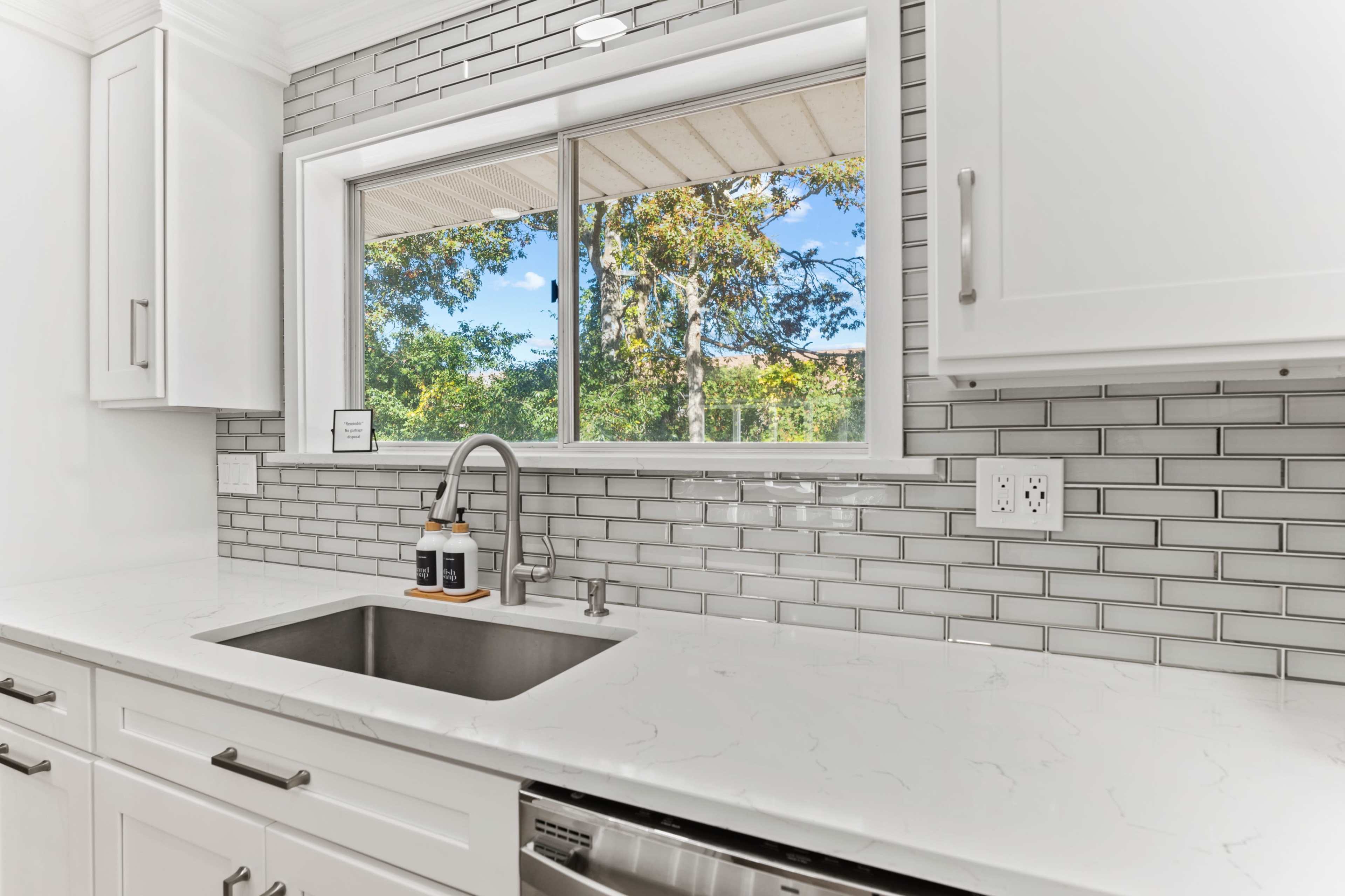 A modern kitchen features a stainless steel sink and a large window overlooking a green outdoor area, with white cabinets and gray backsplash tiles.