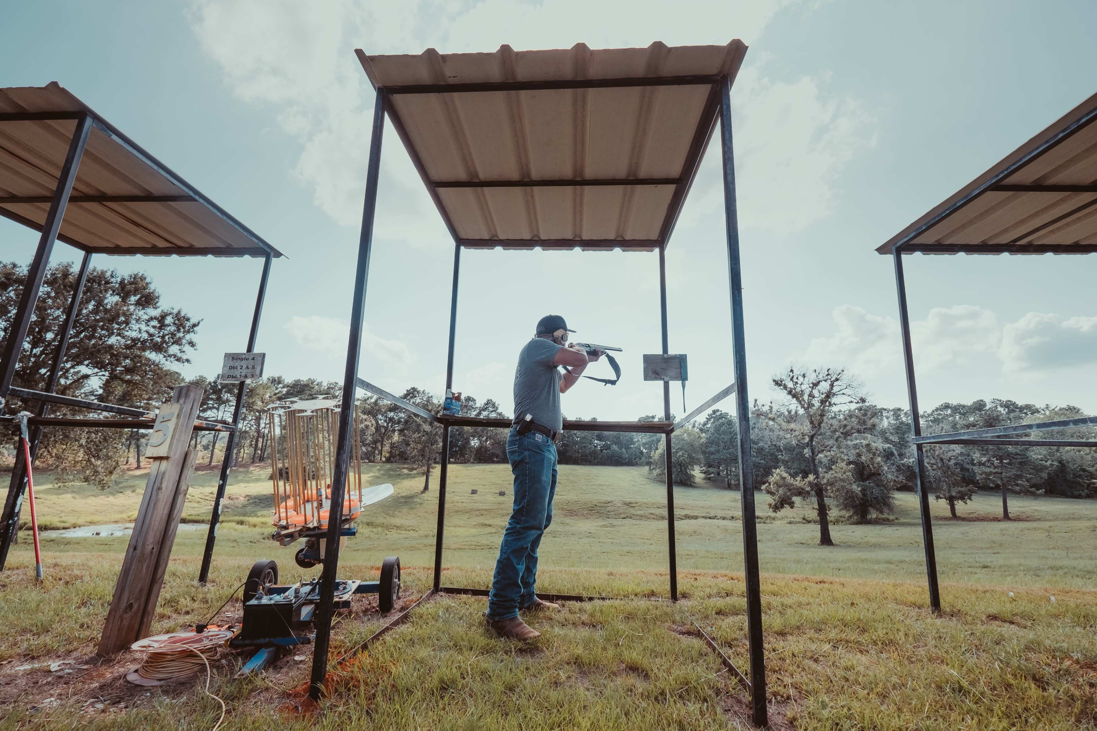A person stands at a shooting station with a shotgun, aiming downrange in a grassy field under a cloudy sky.