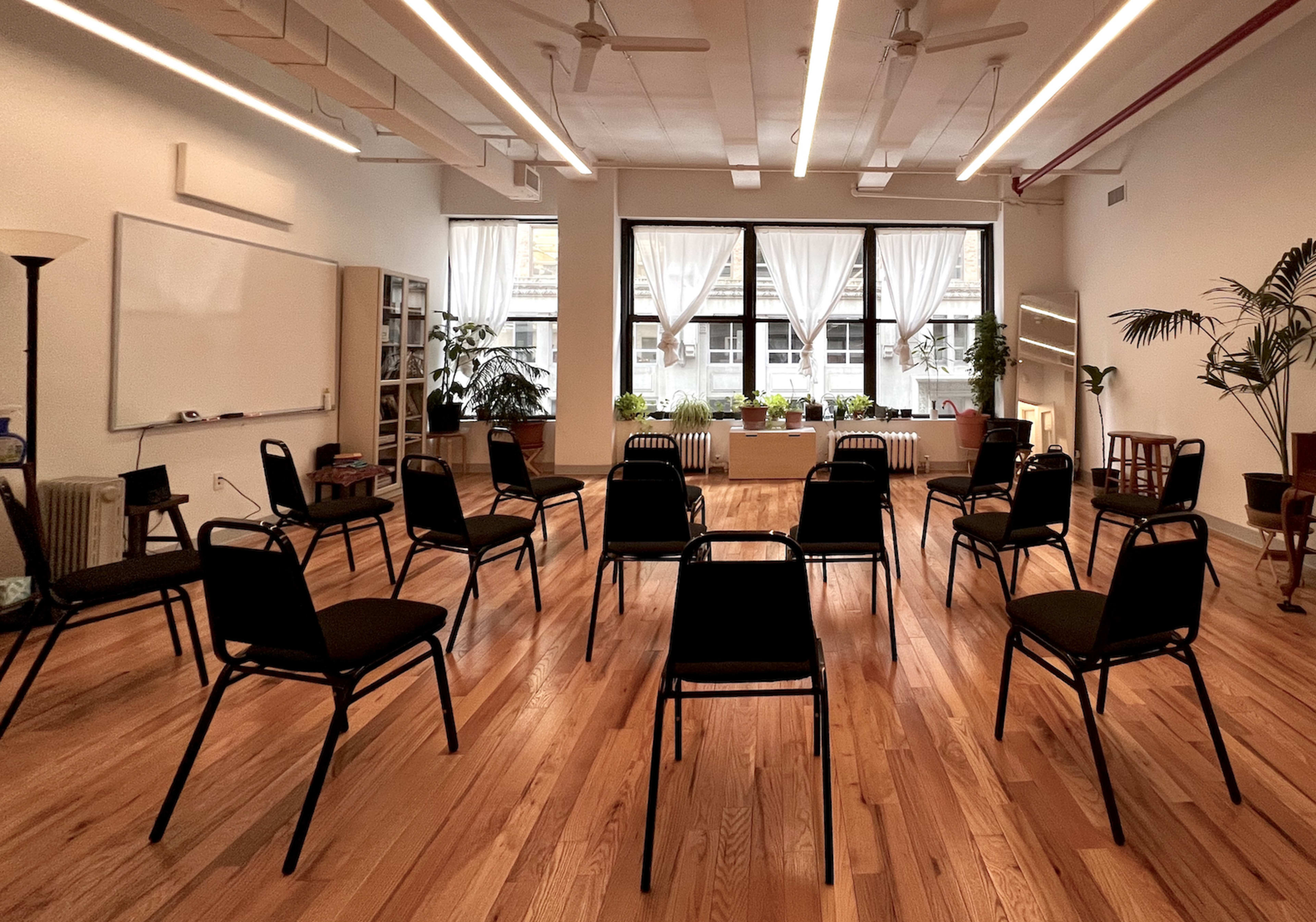 A large, open room with wooden flooring, arranged with chairs in a circular formation around a table, and large windows with plants situated on the sill.