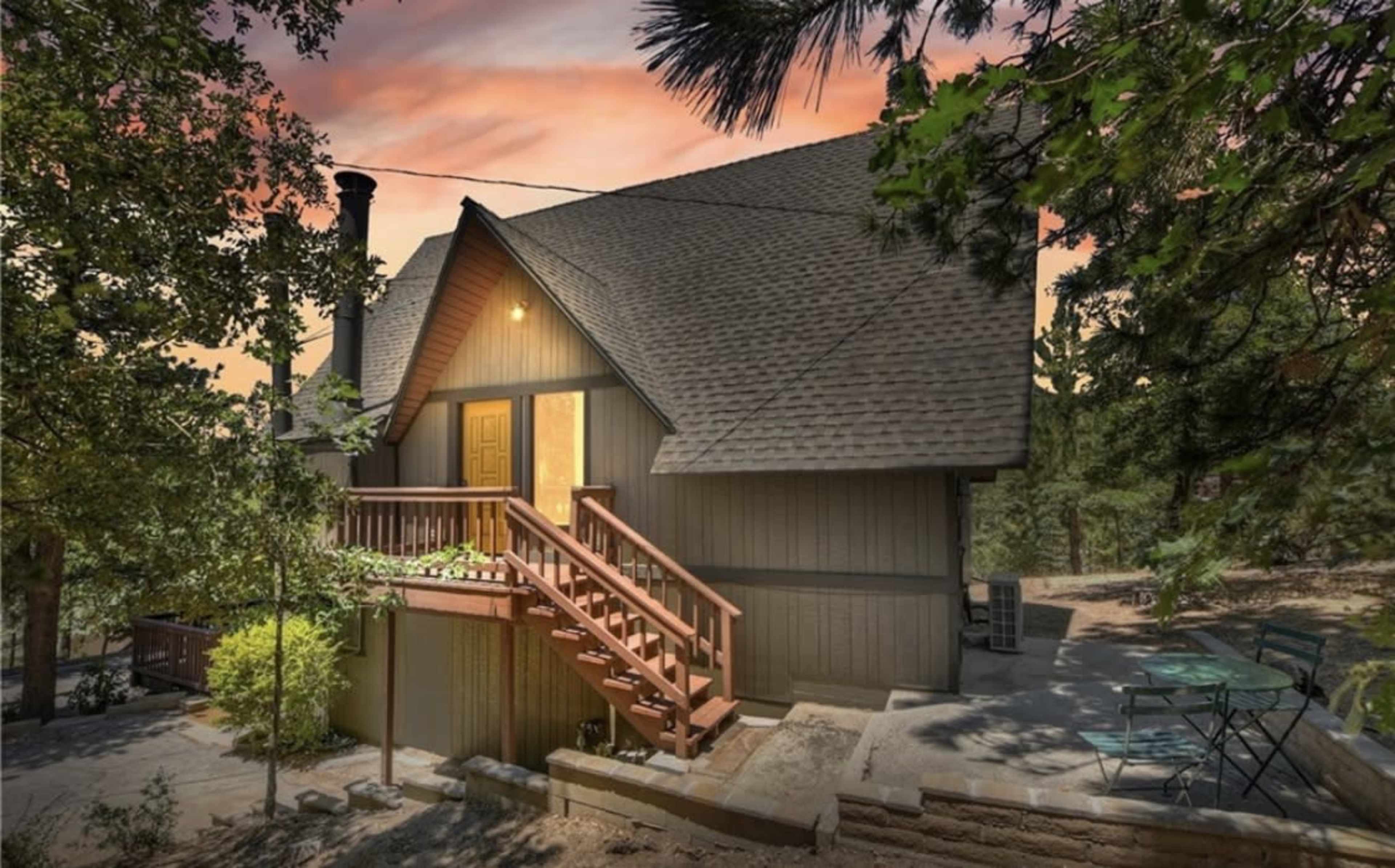 A cabin with a triangular roof, wooden stairs leading to the entrance, and outdoor seating surrounded by trees at sunset.