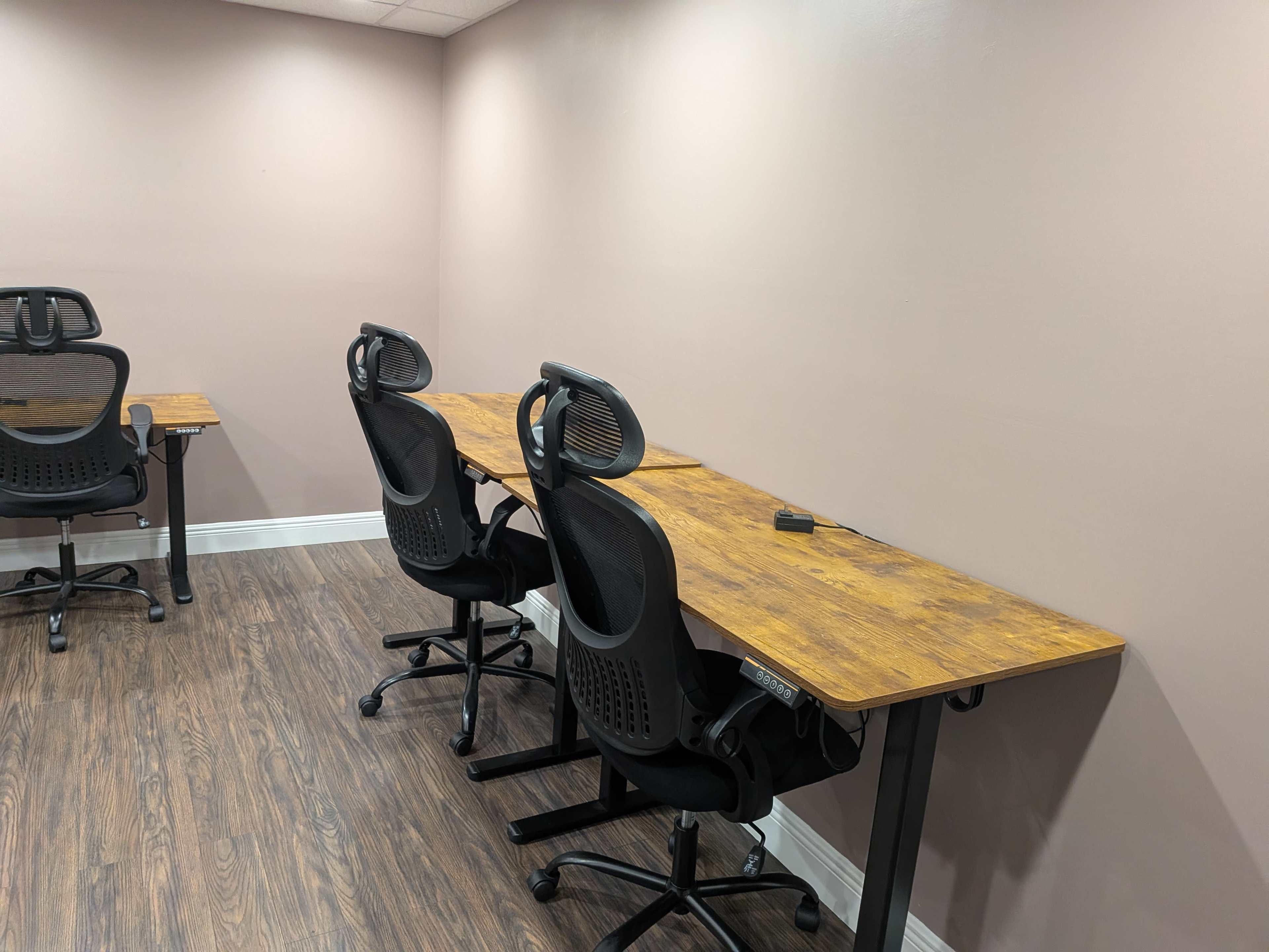 The image shows a sparsely furnished office space with two desks and ergonomic chairs arranged against a light pink wall.