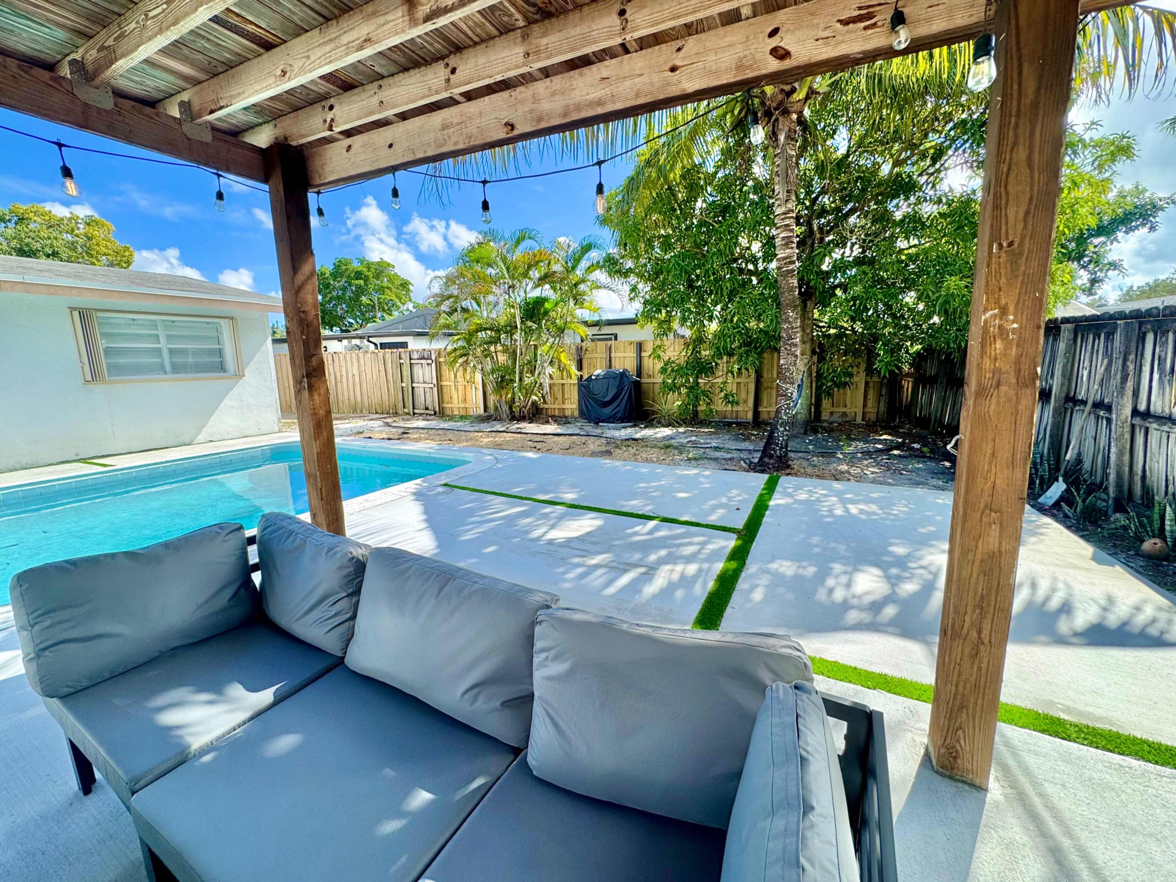 The image shows a covered outdoor area with a gray sofa set, a swimming pool in the background, and a landscaped yard featuring palm trees and a patio.