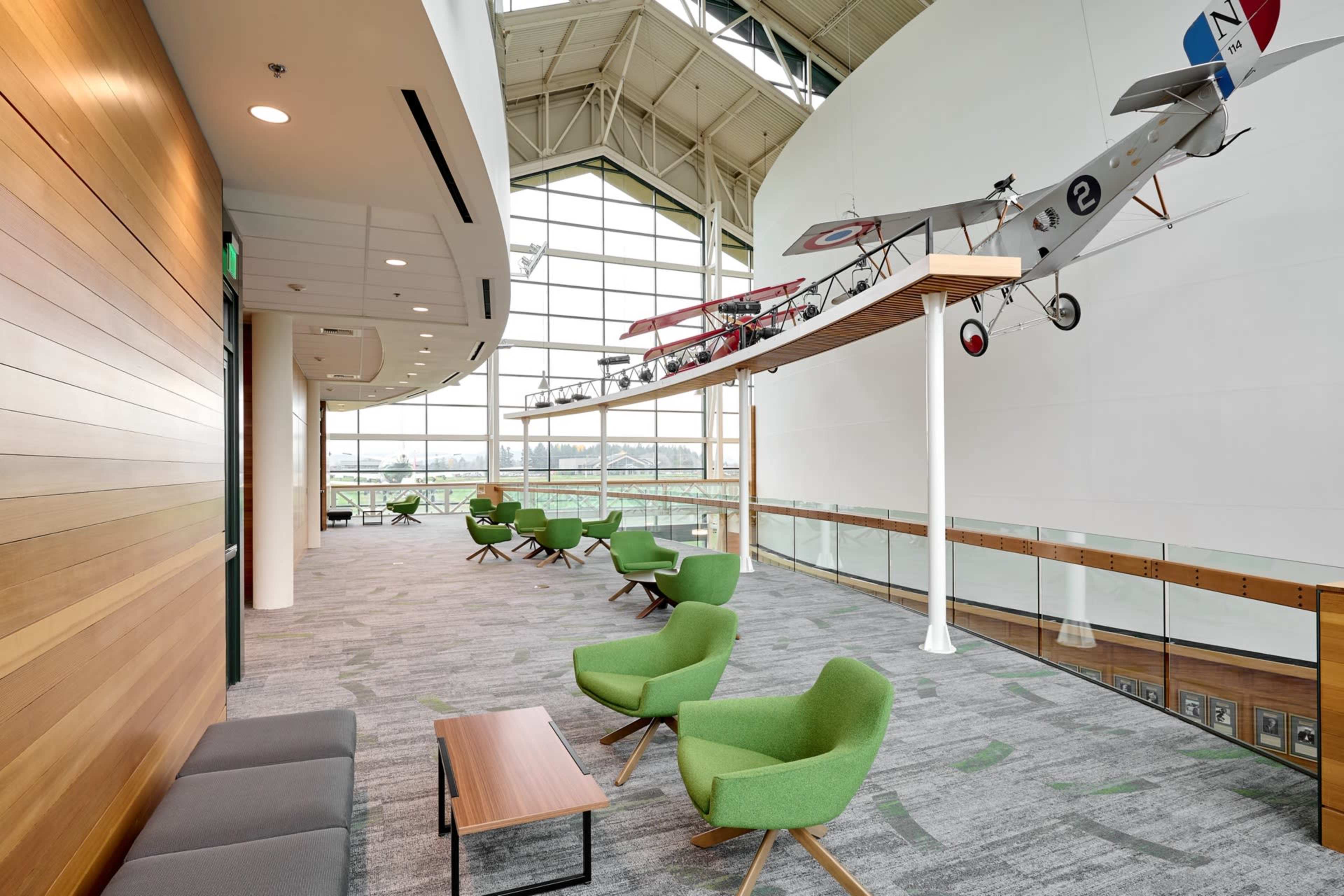 The image depicts a modern indoor space featuring green seating arrangements, wooden paneling, and a vintage airplane suspended from the high ceiling.