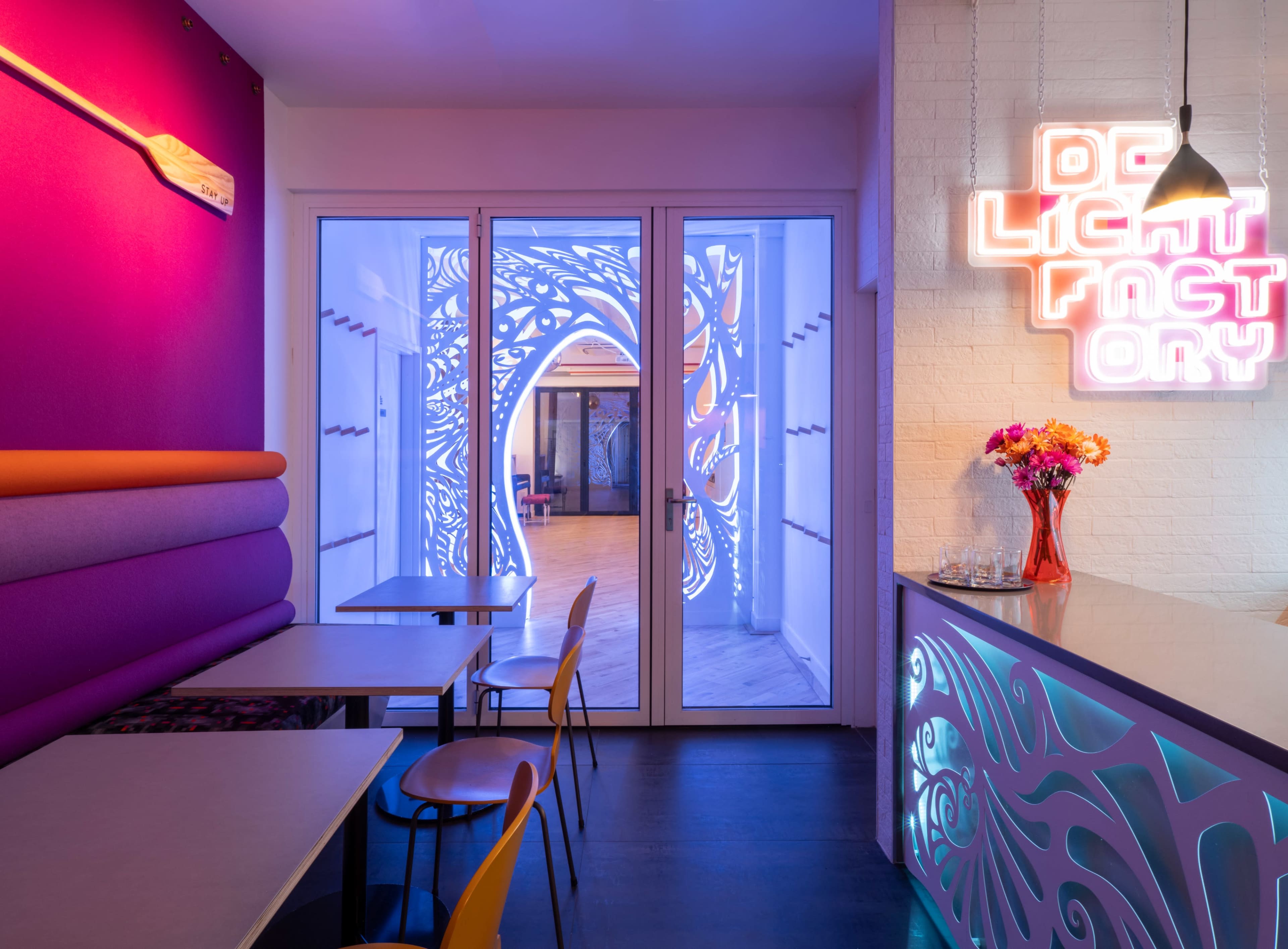 A brightly lit café features colorful walls, modern furniture, and a neon sign alongside a doorway leading to an intricately designed space.