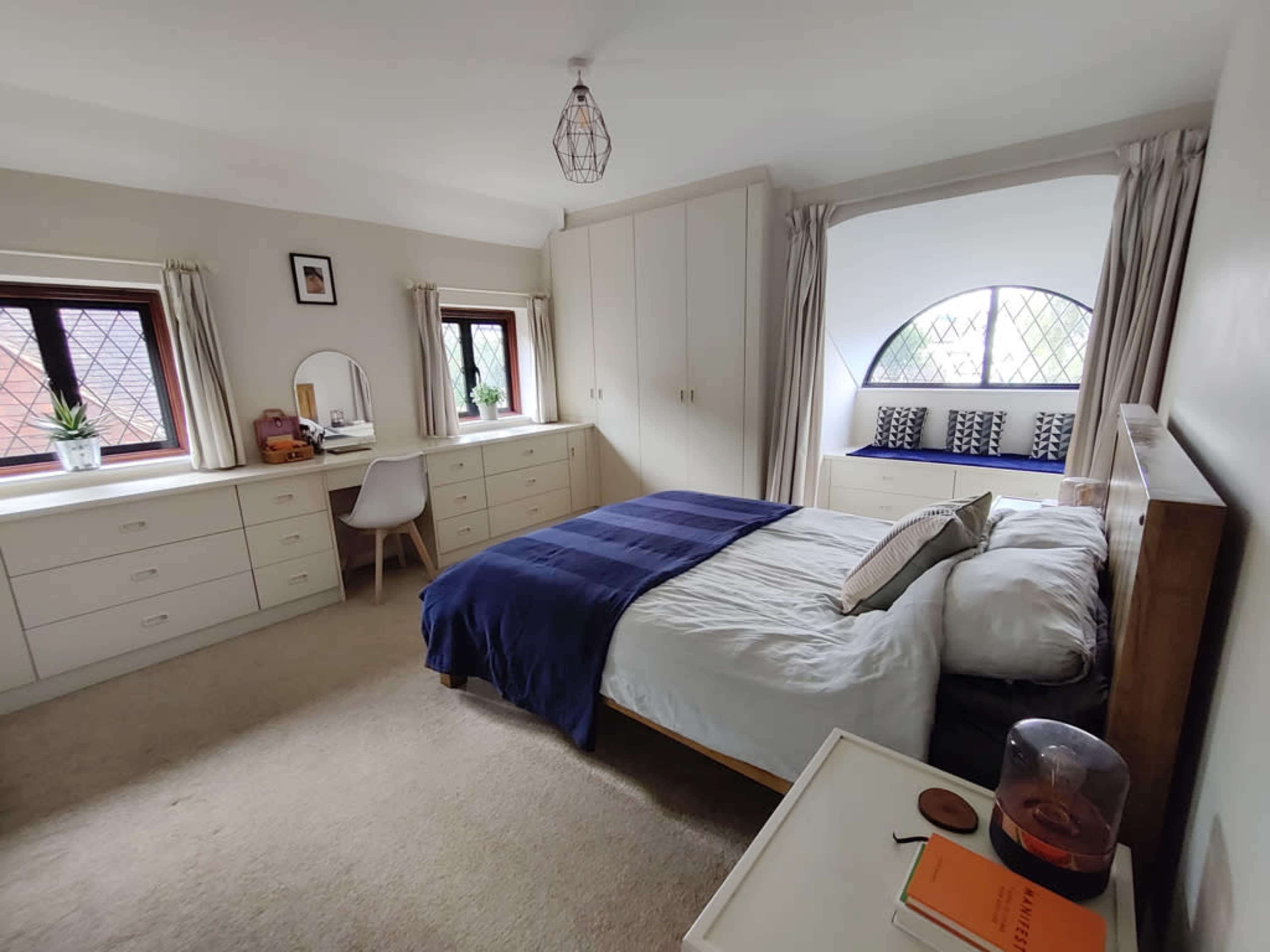 A neatly arranged bedroom featuring a bed with a blue throw blanket, built-in cabinets, a desk with a chair, and a window nook with cushions.
