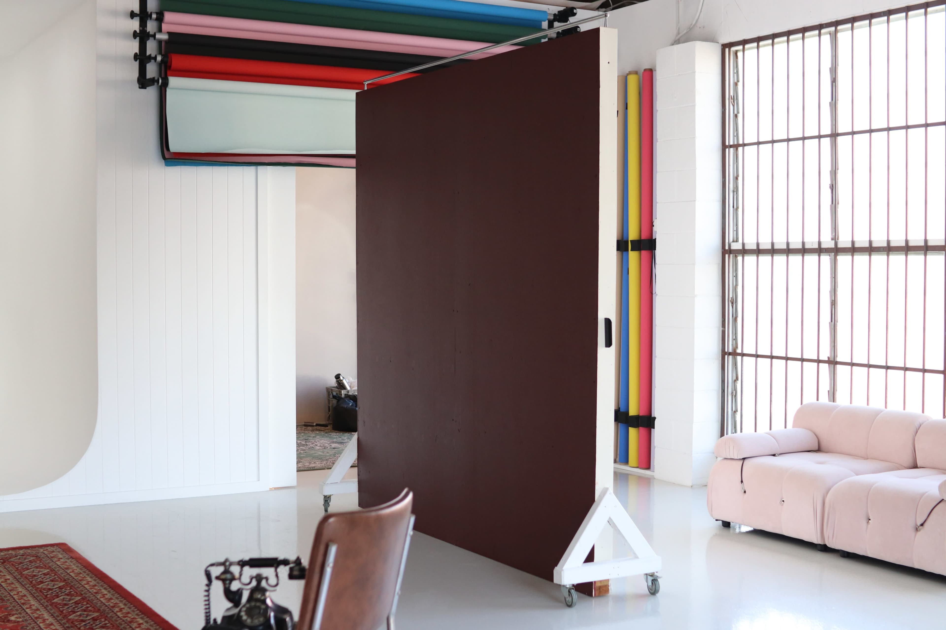 The image shows a photography studio featuring a large brown backdrop on wheels, colorful rolls of seamless paper above, and a light-colored sofa in the corner.