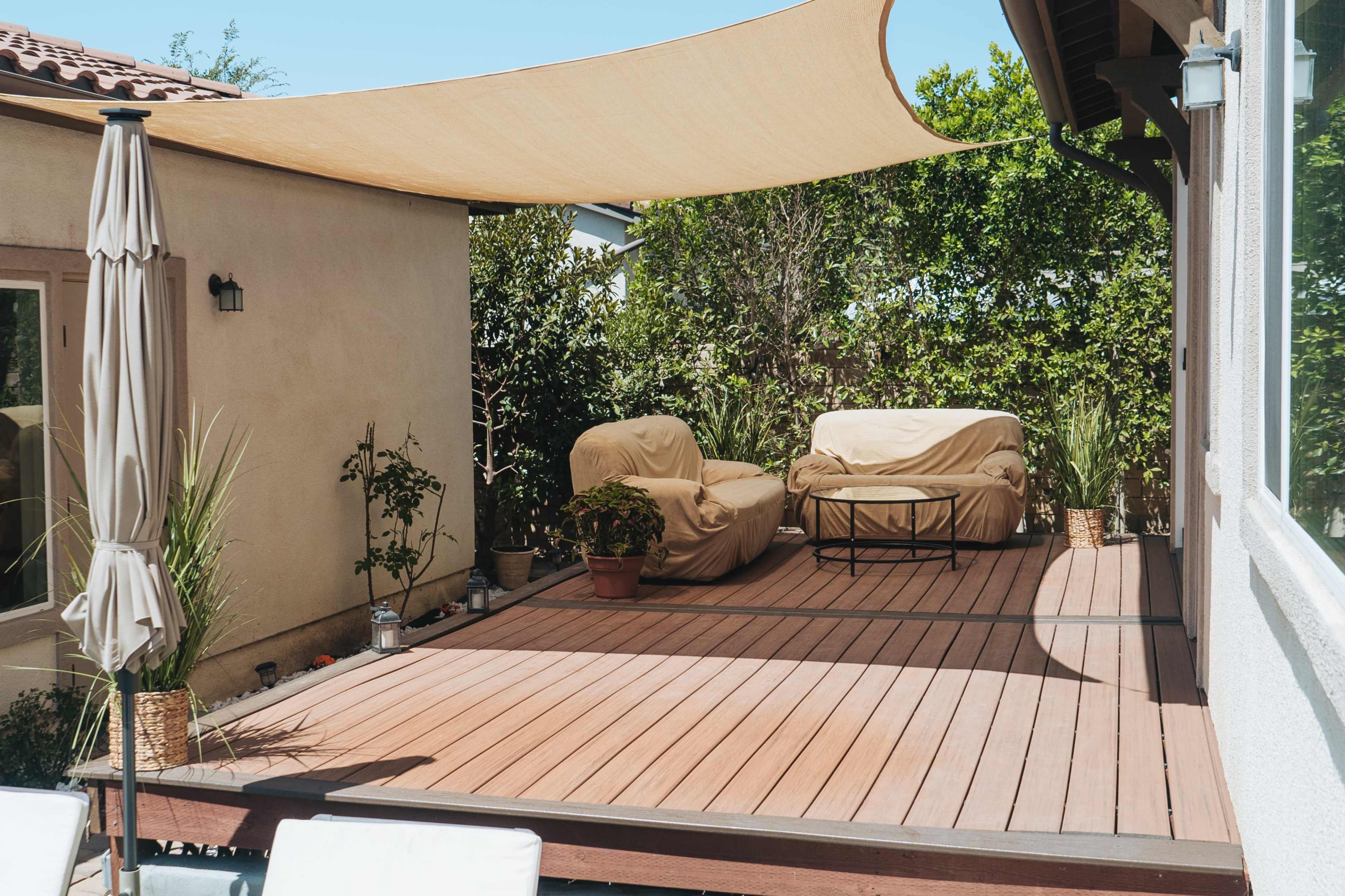 A wooden deck with a table and two covered chairs is shaded by a fabric awning next to a house surrounded by greenery.