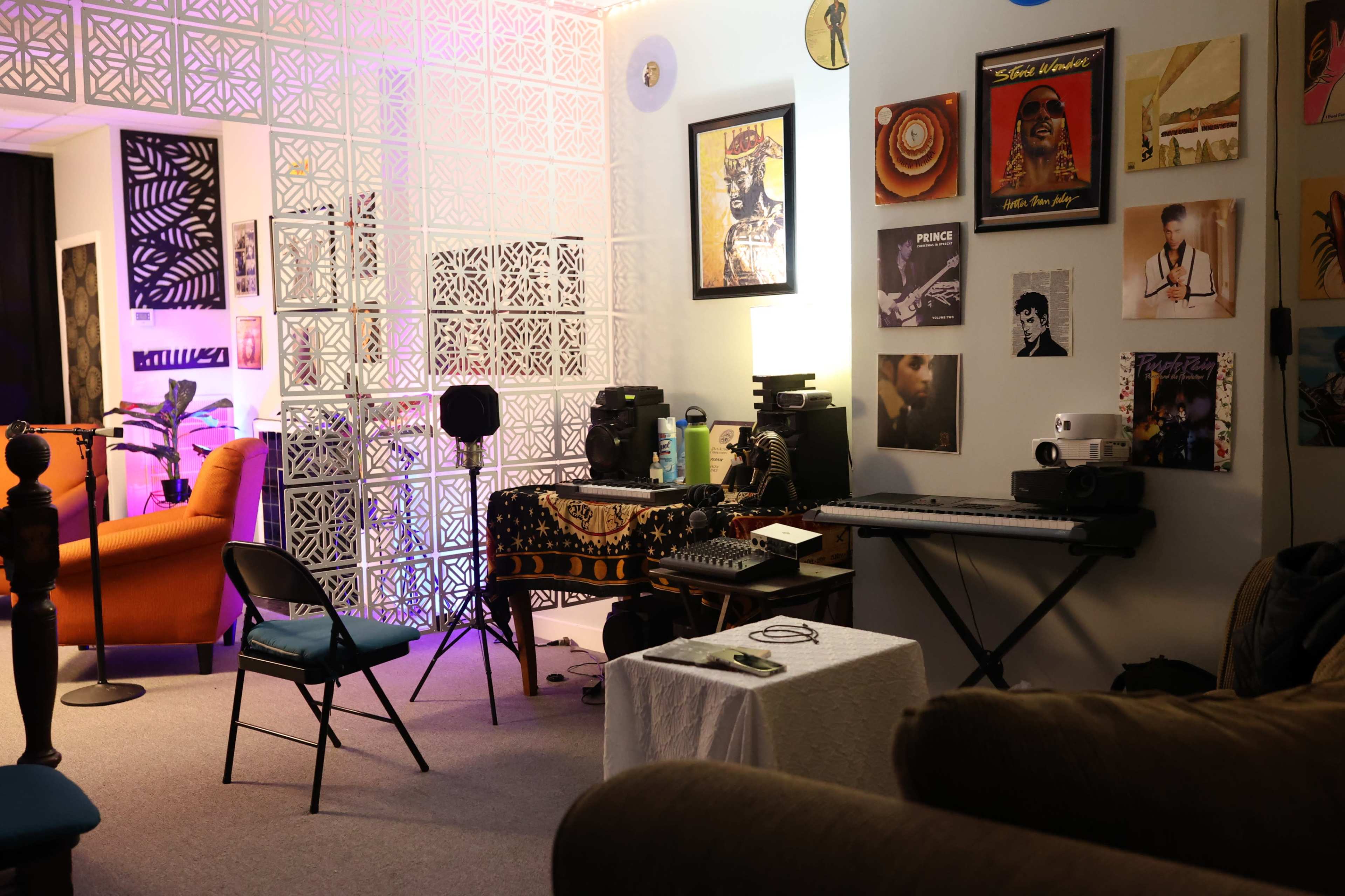A cozy music studio with a keyboard setup, speakers, and framed records on the walls, accompanied by chairs and a decorative screen.