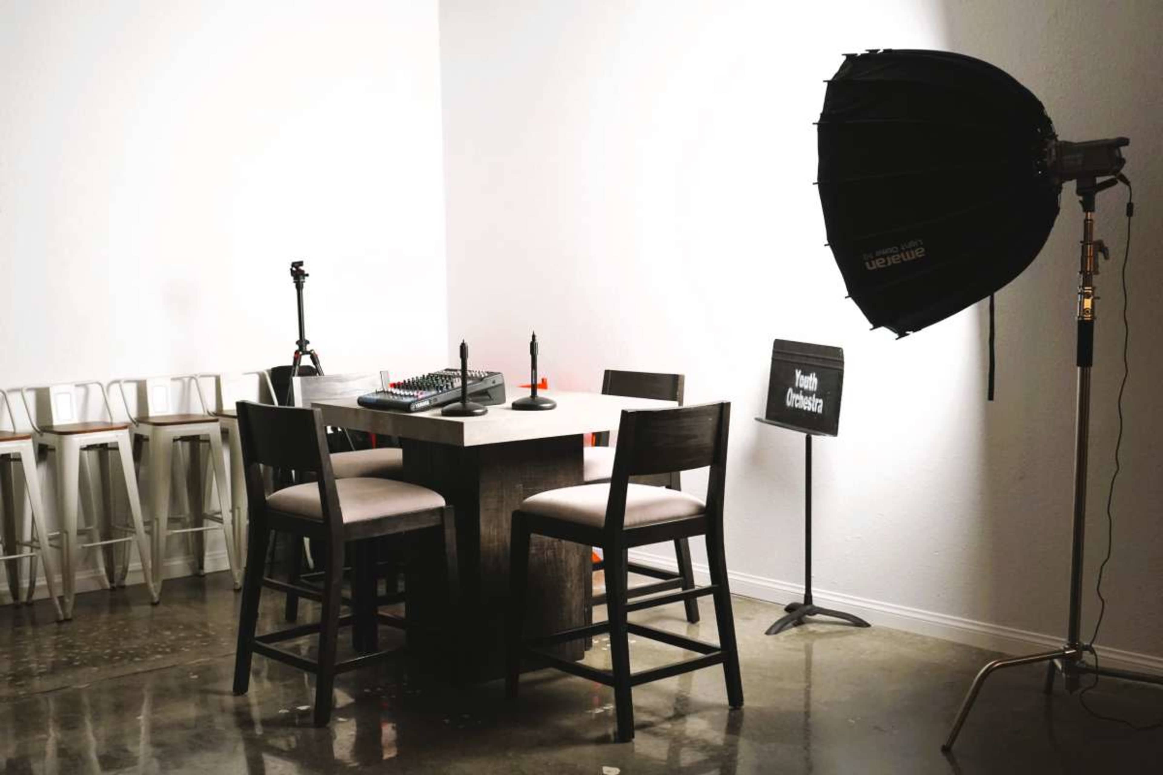 A small recording studio setup features a table with audio equipment, four chairs, and a large light umbrella in the corner.