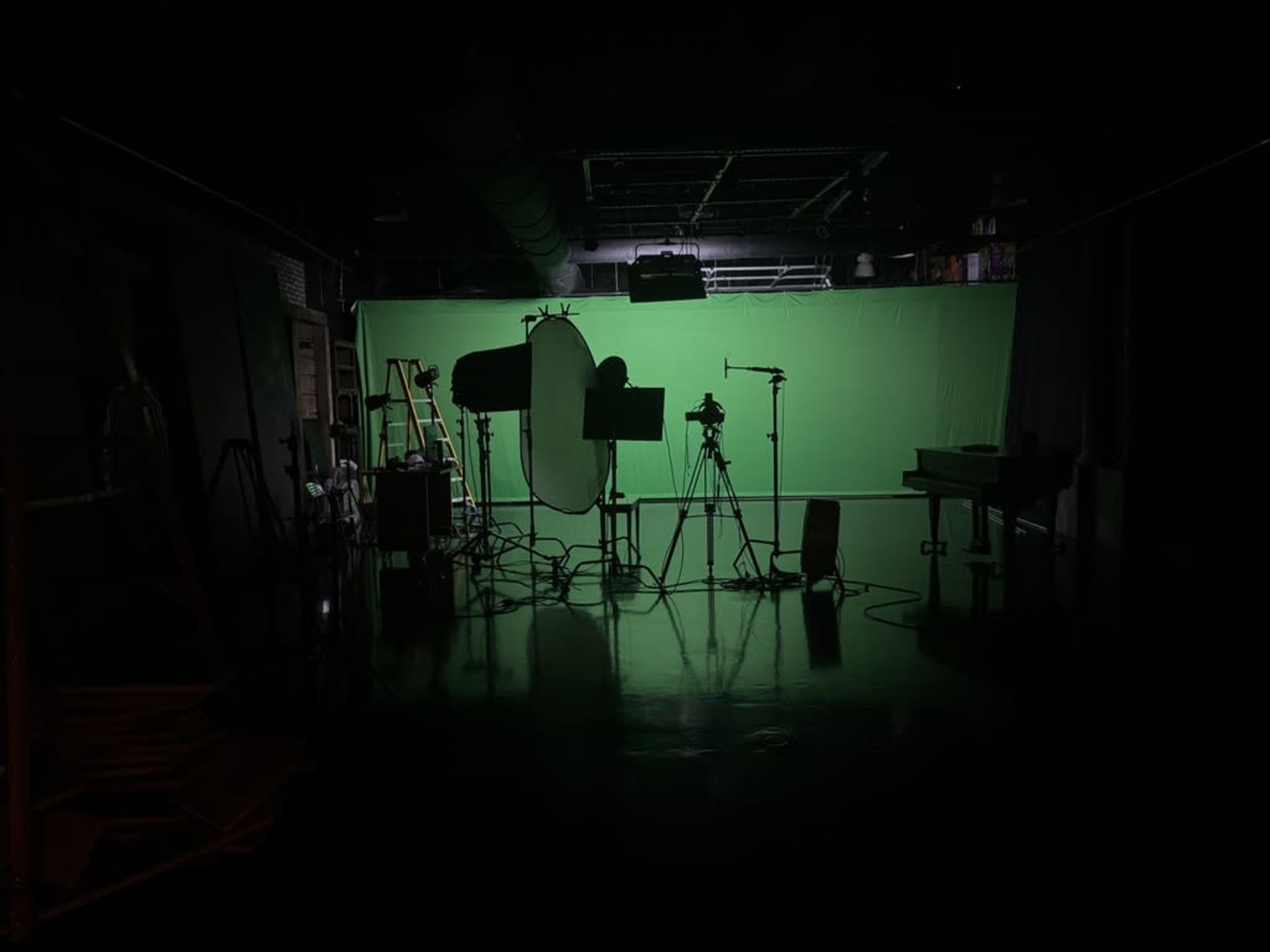 A dimly lit studio features various equipment and a large green screen as the backdrop.