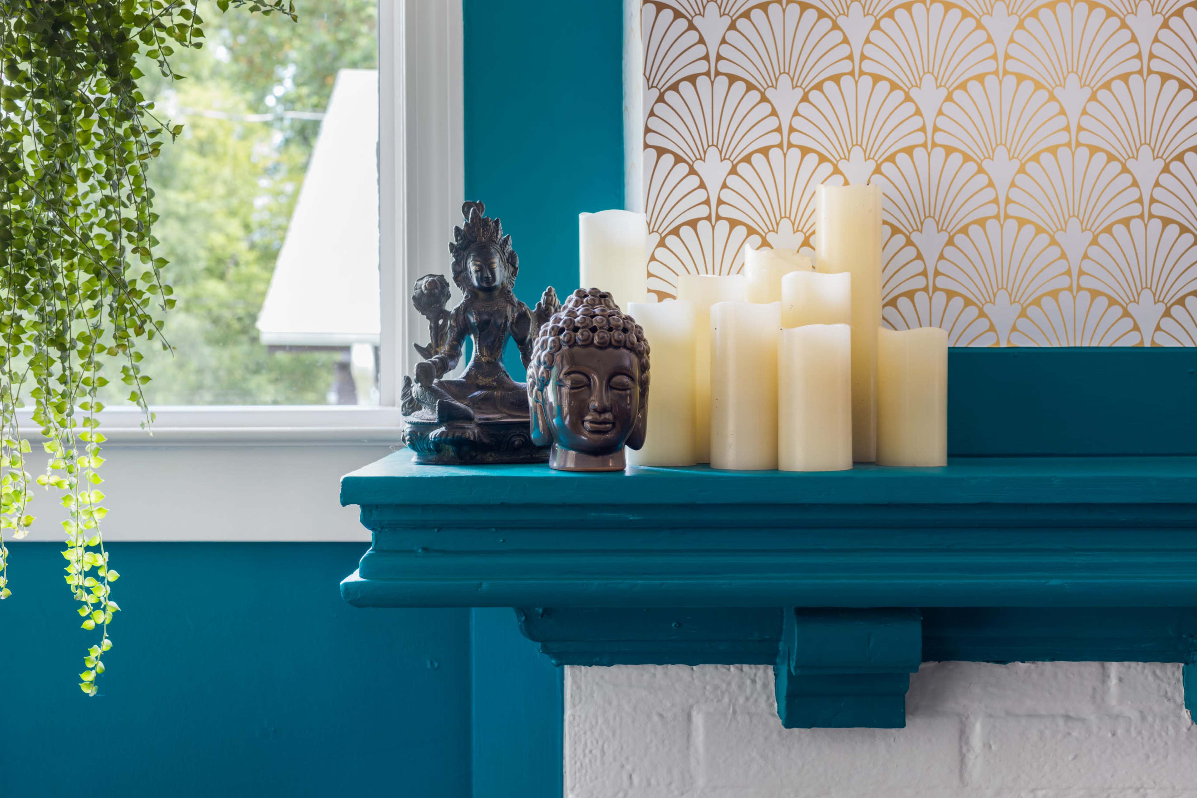 A wooden shelf features decorative items, including a Buddha statue, a small figure of a deity, and several LED candles against a teal wall.