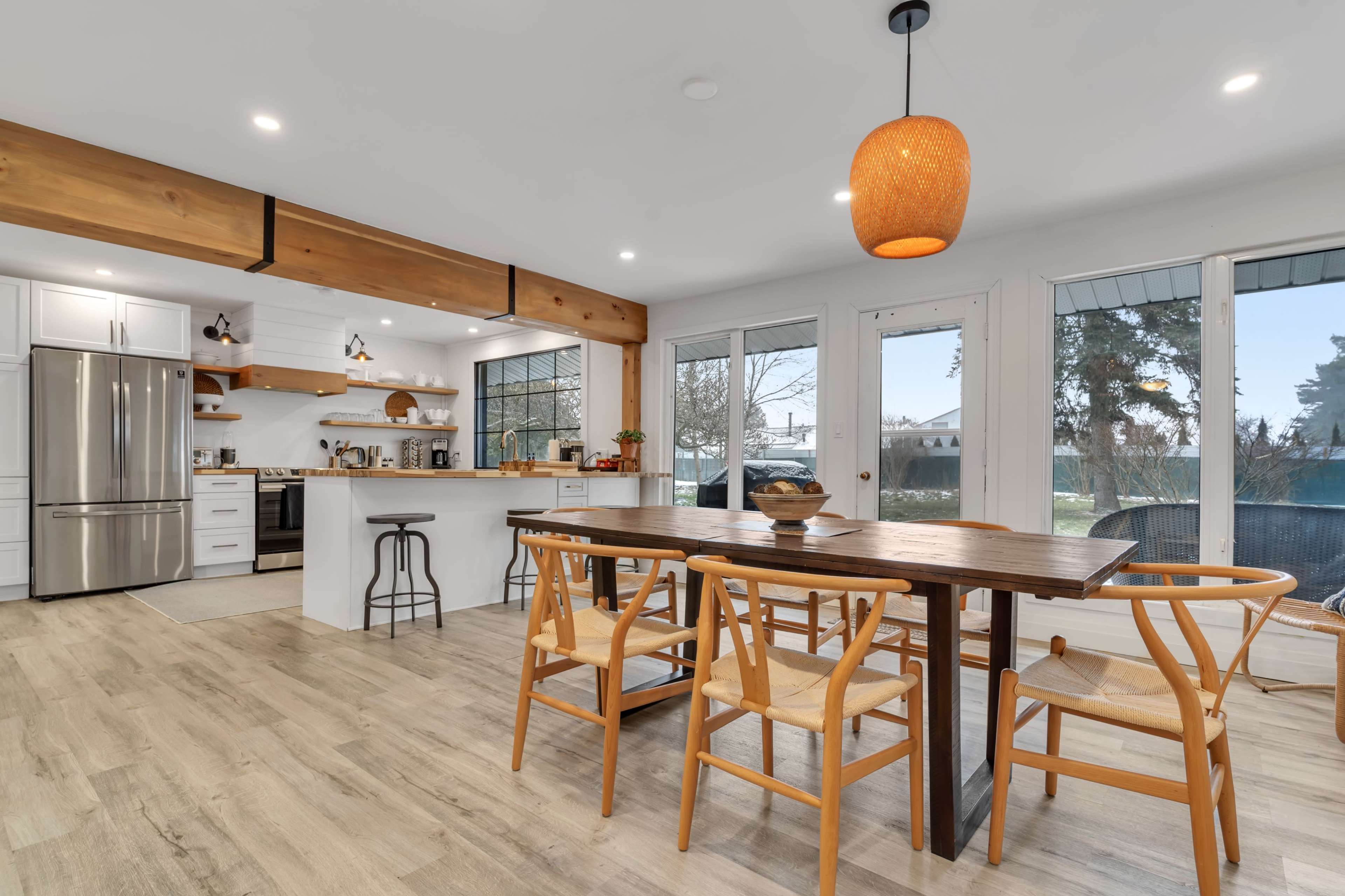 A modern kitchen features a wooden dining table surrounded by chairs, with a large window illuminating stainless steel appliances and open shelving.