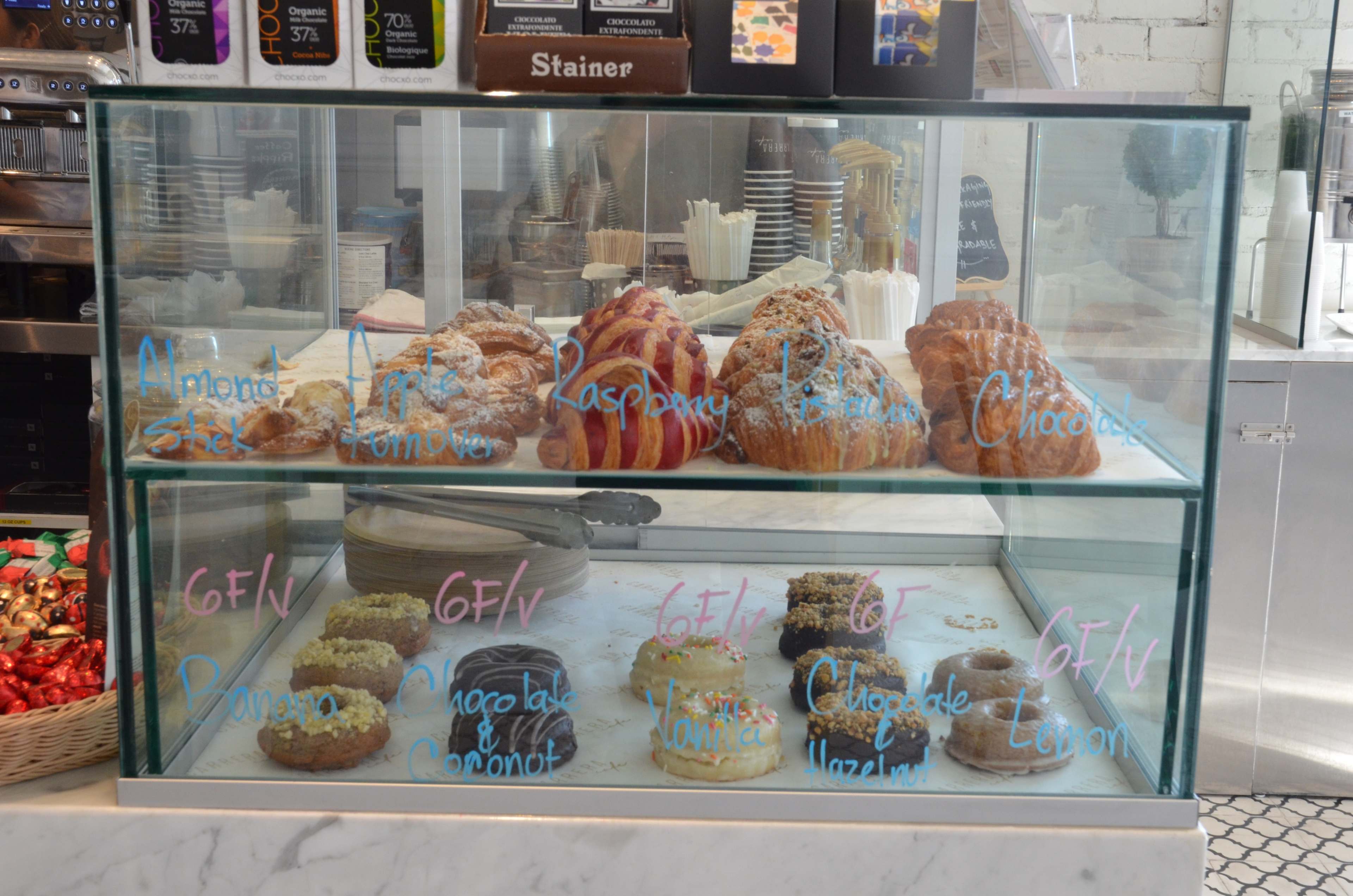 A glass display case showcases various pastries, including almond croissants, raspberry danishes, chocolate hazelnut treats, and assorted donuts.