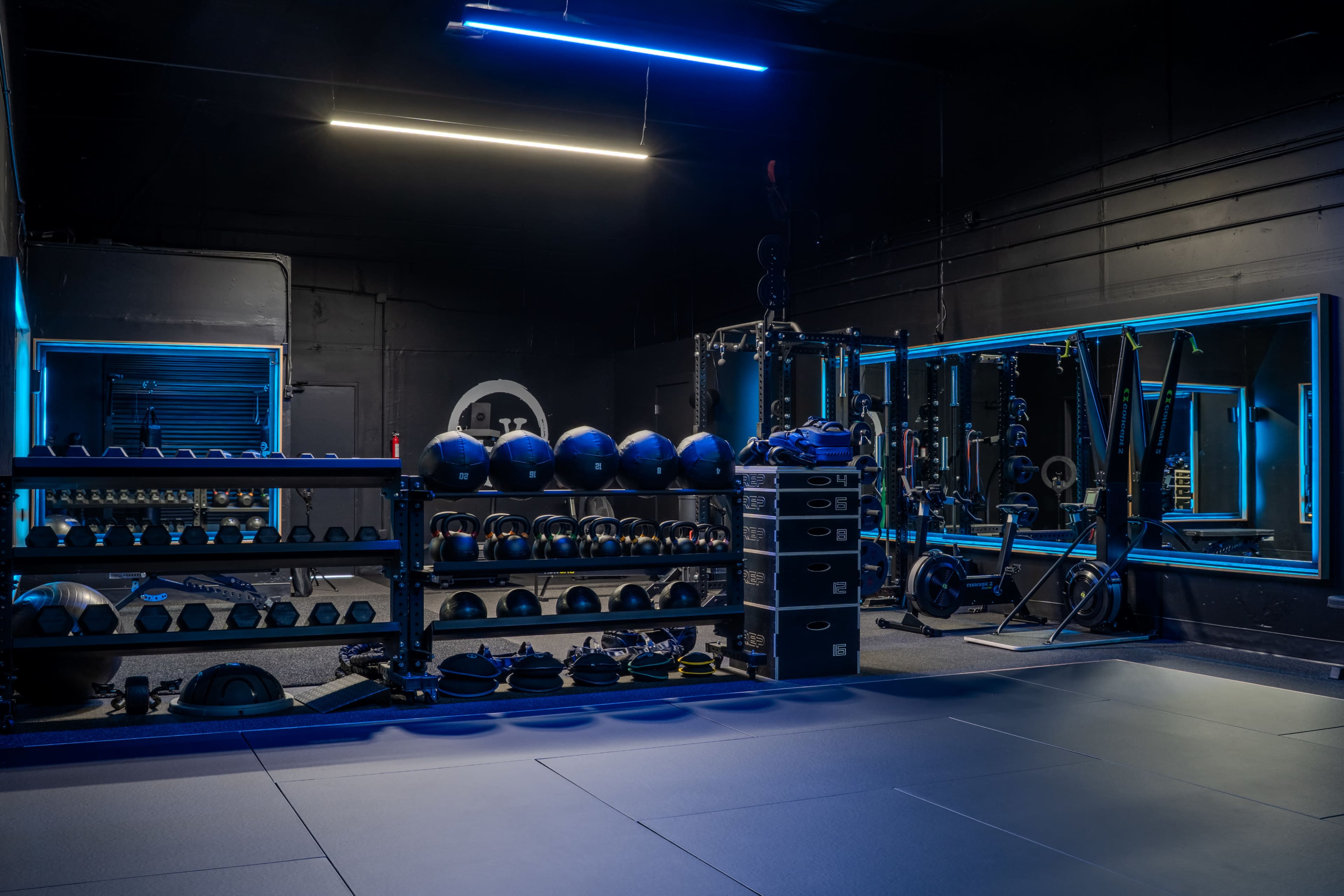 The image shows a modern gym interior with various fitness equipment, including weights and exercise mats, illuminated by blue lights.