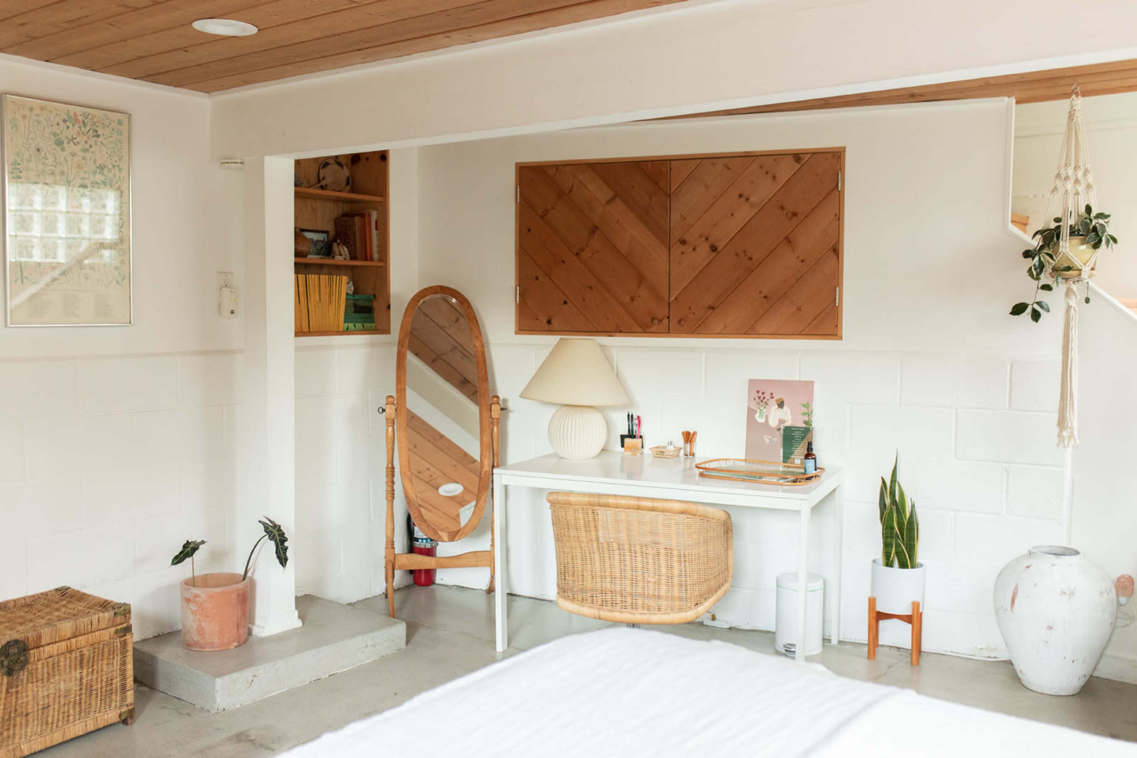 The image shows a minimalist bedroom featuring a desk with a lamp, a mirror, decorative plants, and wooden shelves against a white wall.