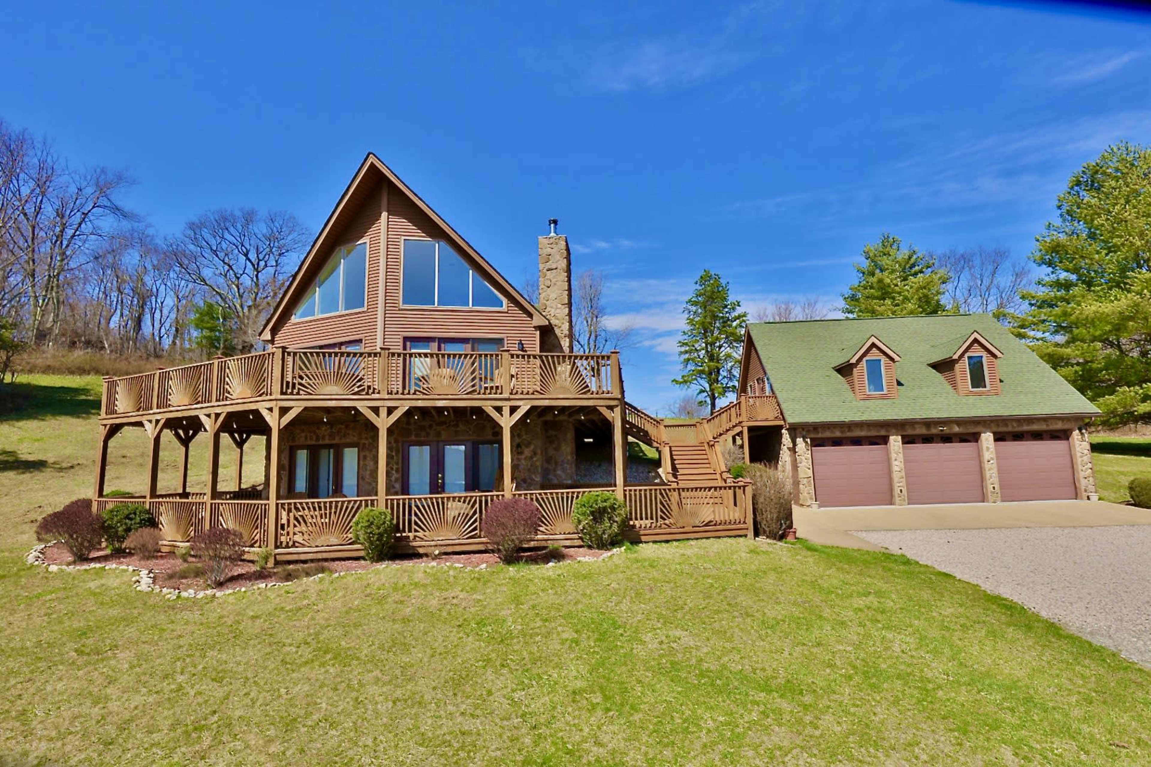 A multi-level house with large windows and a wraparound deck is situated on a grassy hillside, accompanied by a two-car garage.