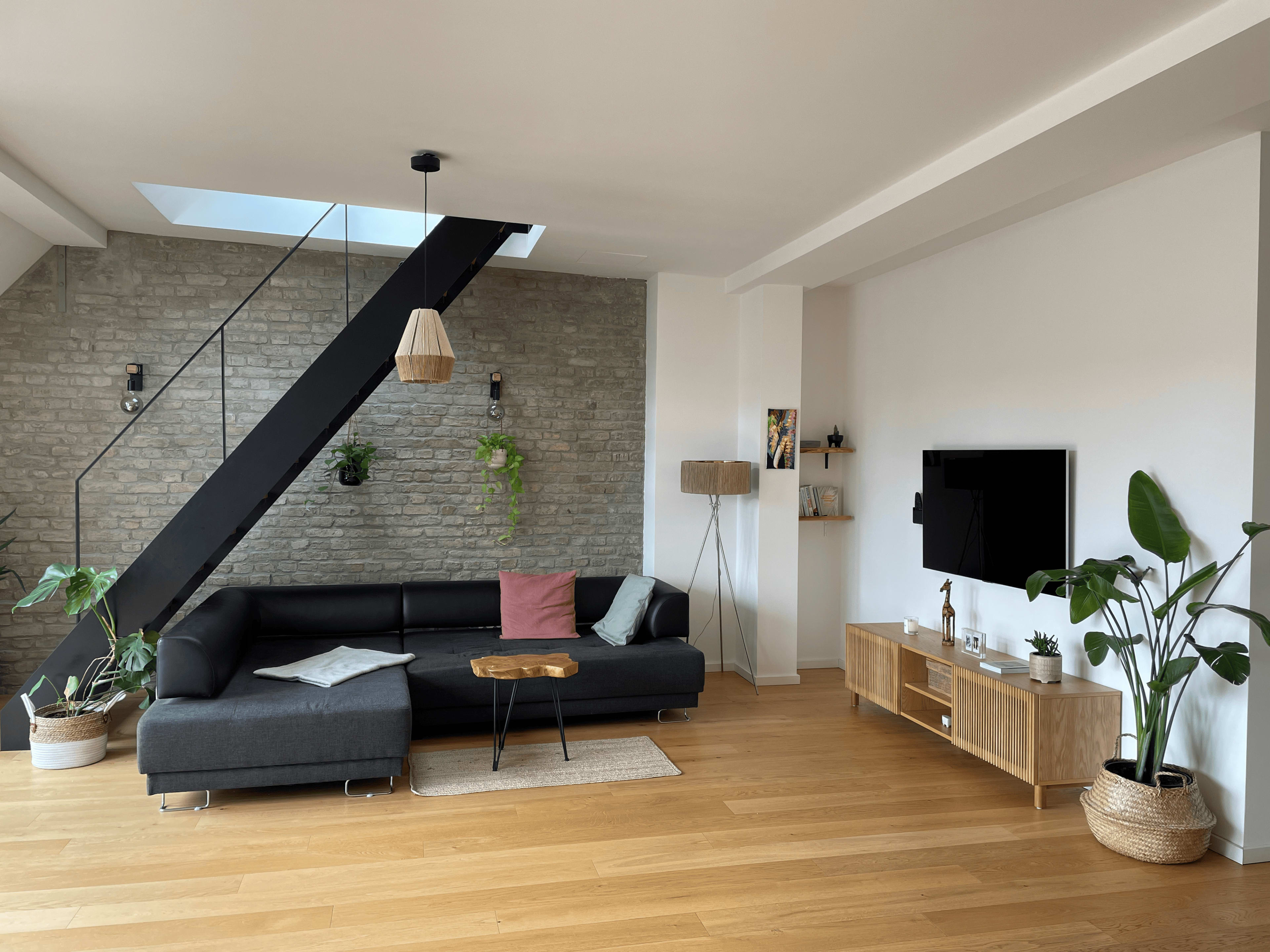 A modern living room featuring a black sectional sofa, a wooden coffee table, a television mounted on a wall, and plants in decorative pots.