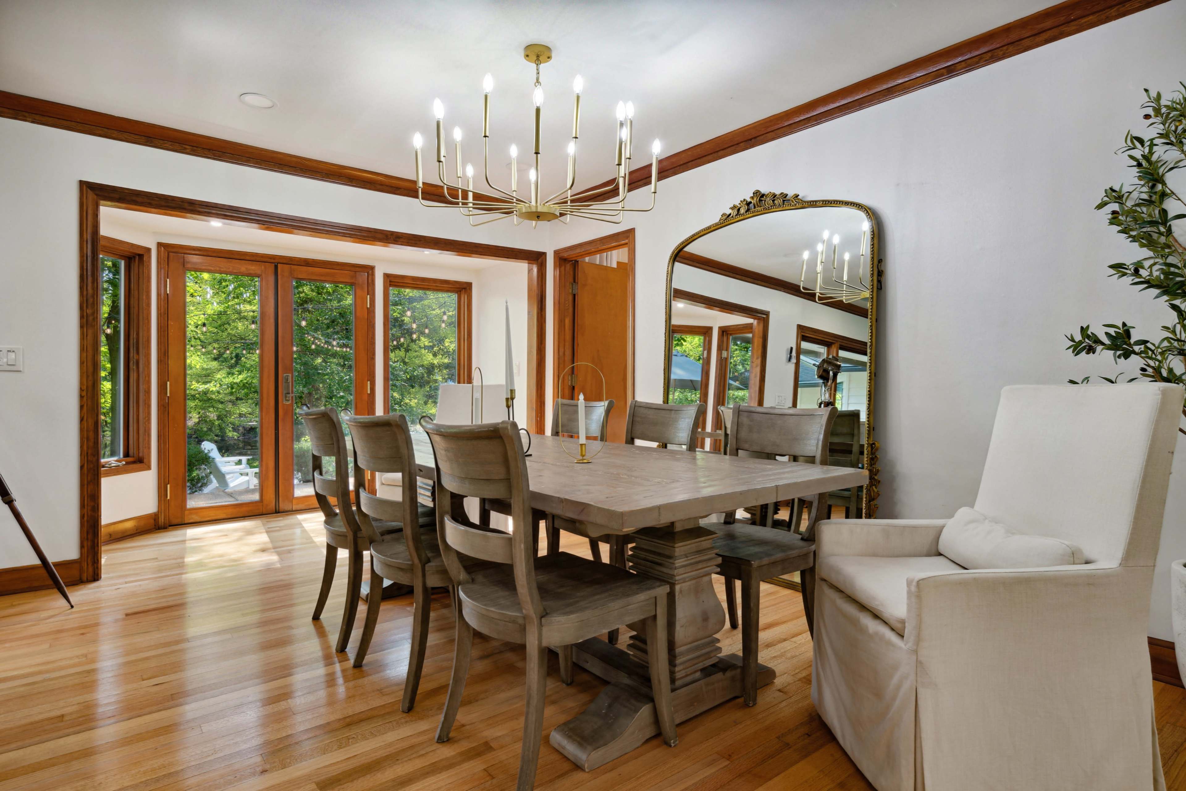 A modern dining room features a large wooden table surrounded by ten chairs, with a chandelier overhead and large windows offering a view of greenery outside.