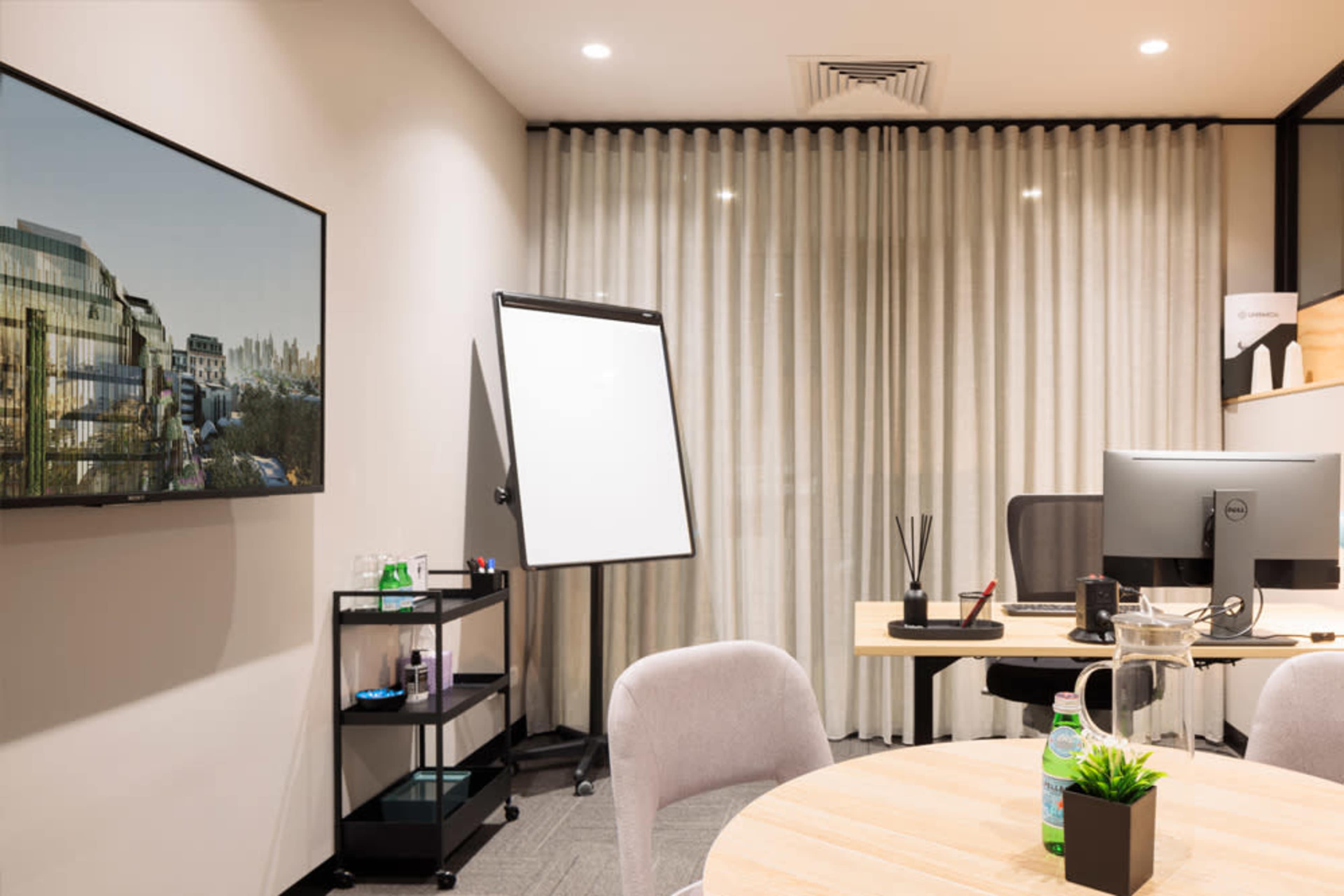 An office meeting room features a large screen, a whiteboard on an easel, a desk with a computer, and a round table with a small plant.