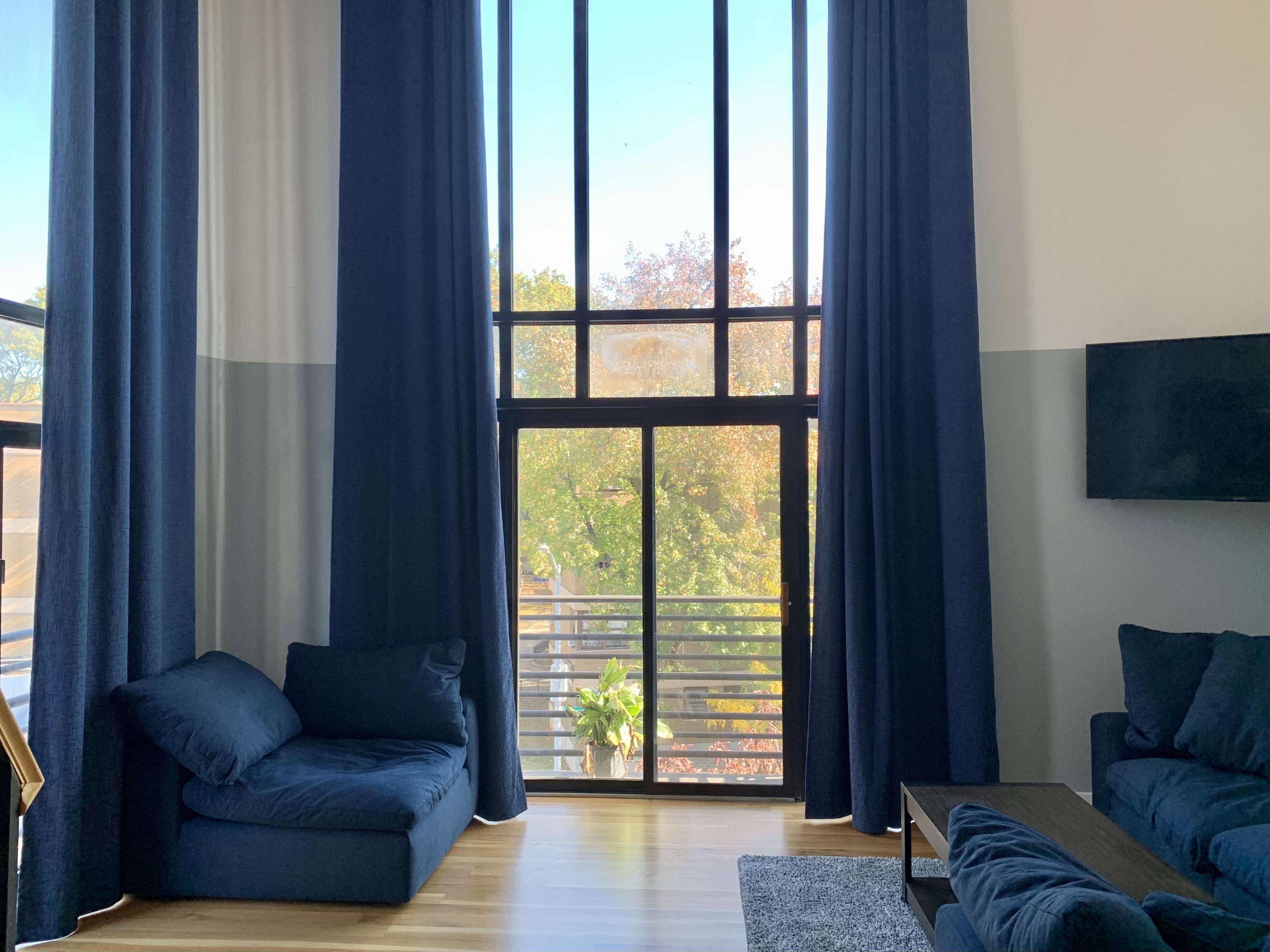 A modern living room features large windows with dark curtains, showcasing a view of trees outside, and includes a seating area with blue couches and a wooden coffee table.