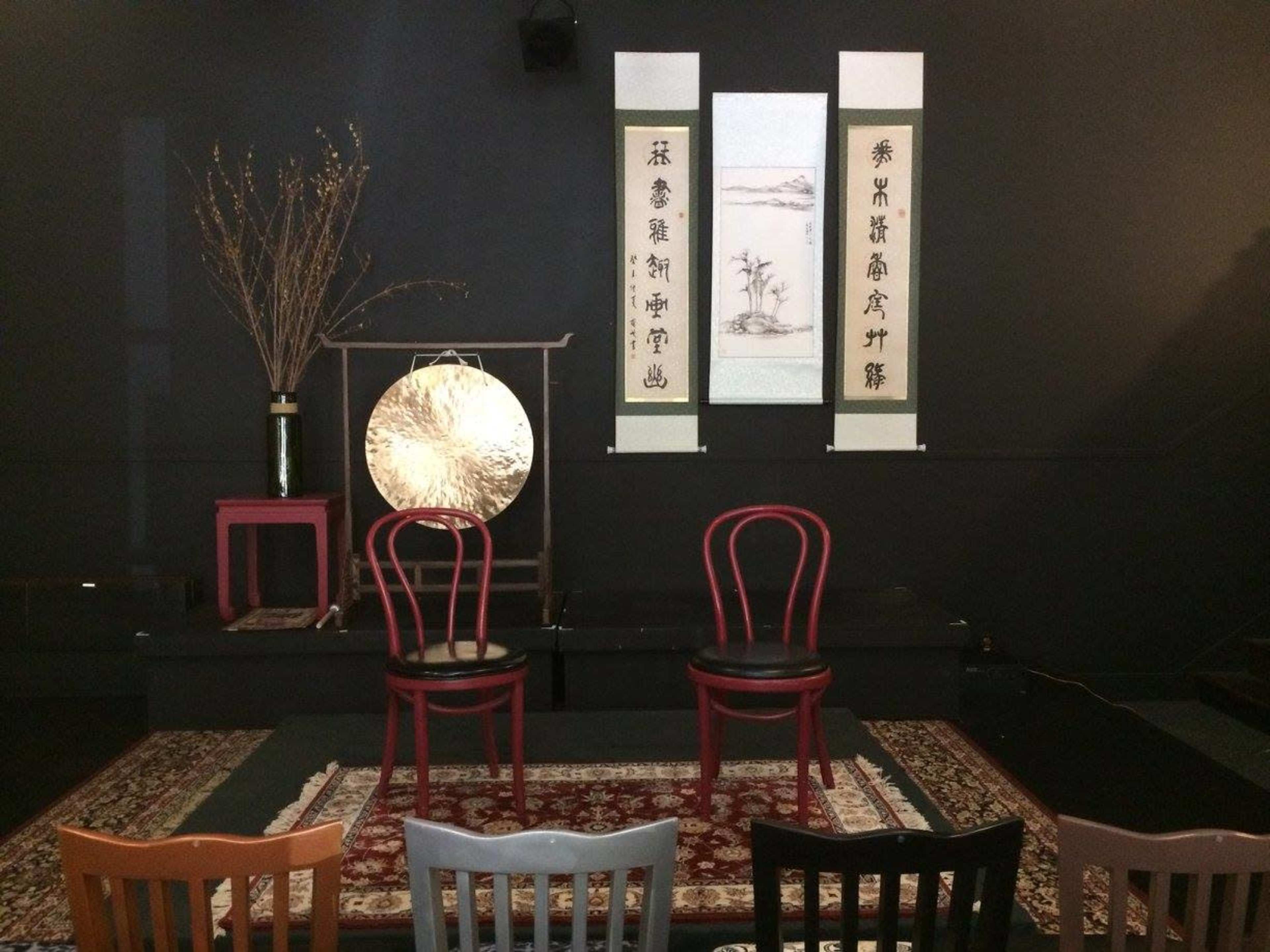 A stage setup with two red chairs, a large decorative gong, and hanging artwork on a black wall, all arranged on a patterned rug.