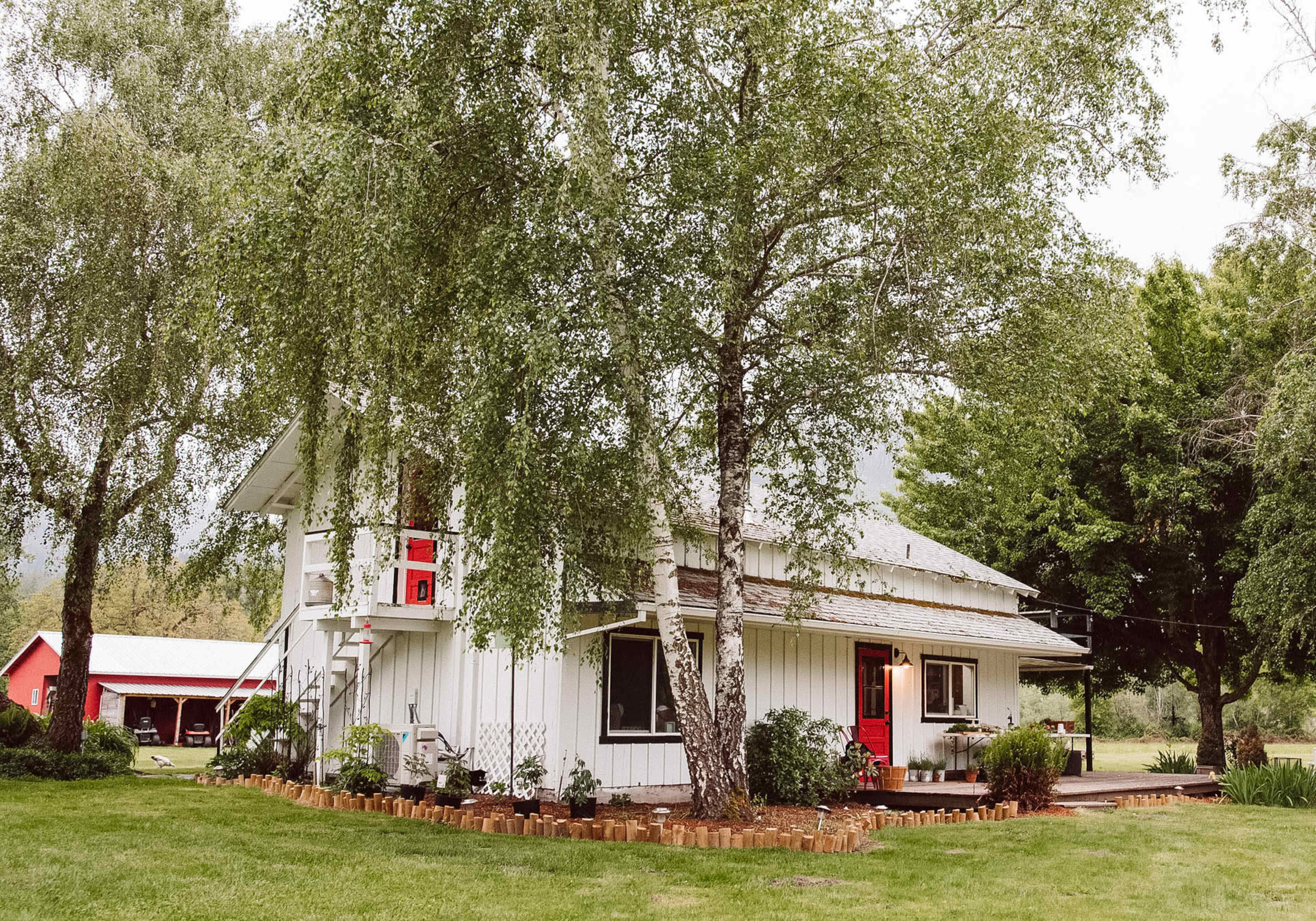 A white two-story house with red accents is surrounded by greenery and a well-maintained lawn.