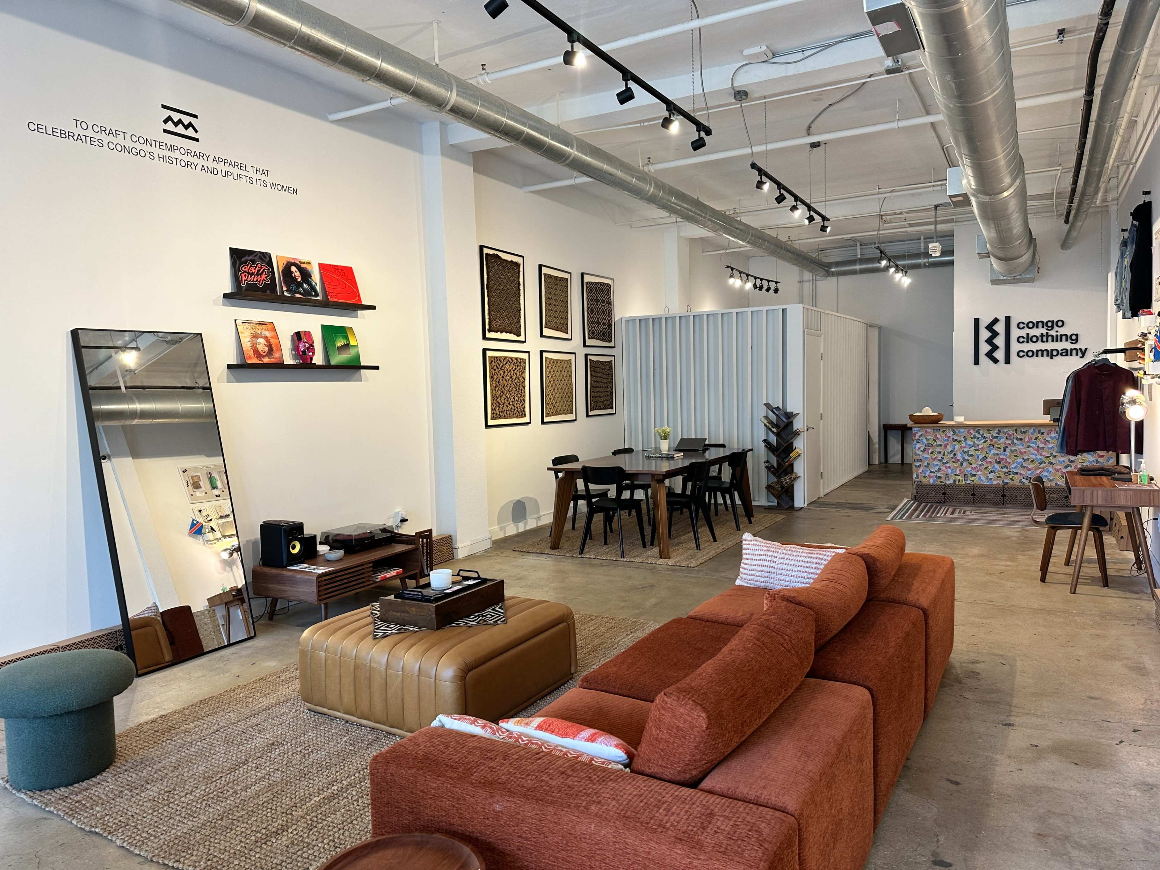 The image shows a modern retail space with a sofa, a dining table, and various artworks displayed on the walls.