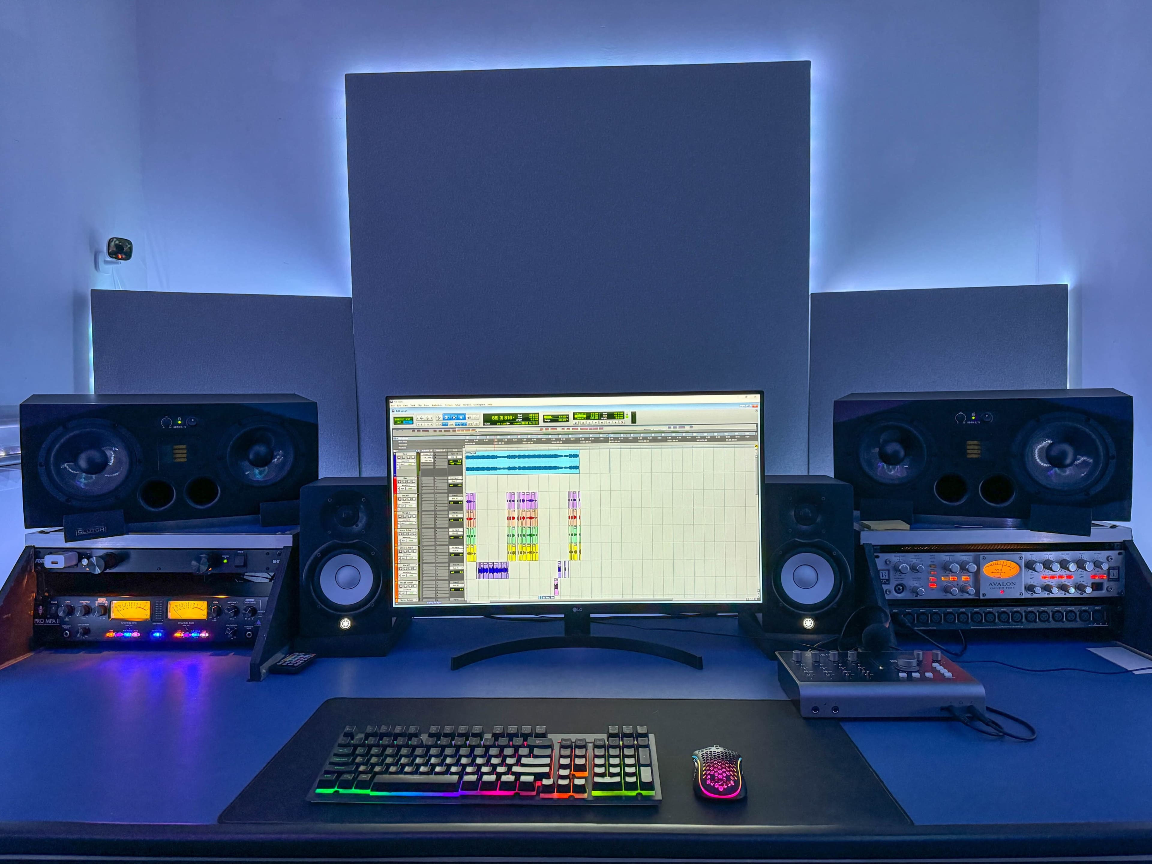 The image shows a modern music production studio with a central computer monitor displaying a digital audio workstation, flanked by studio monitors and various audio equipment.