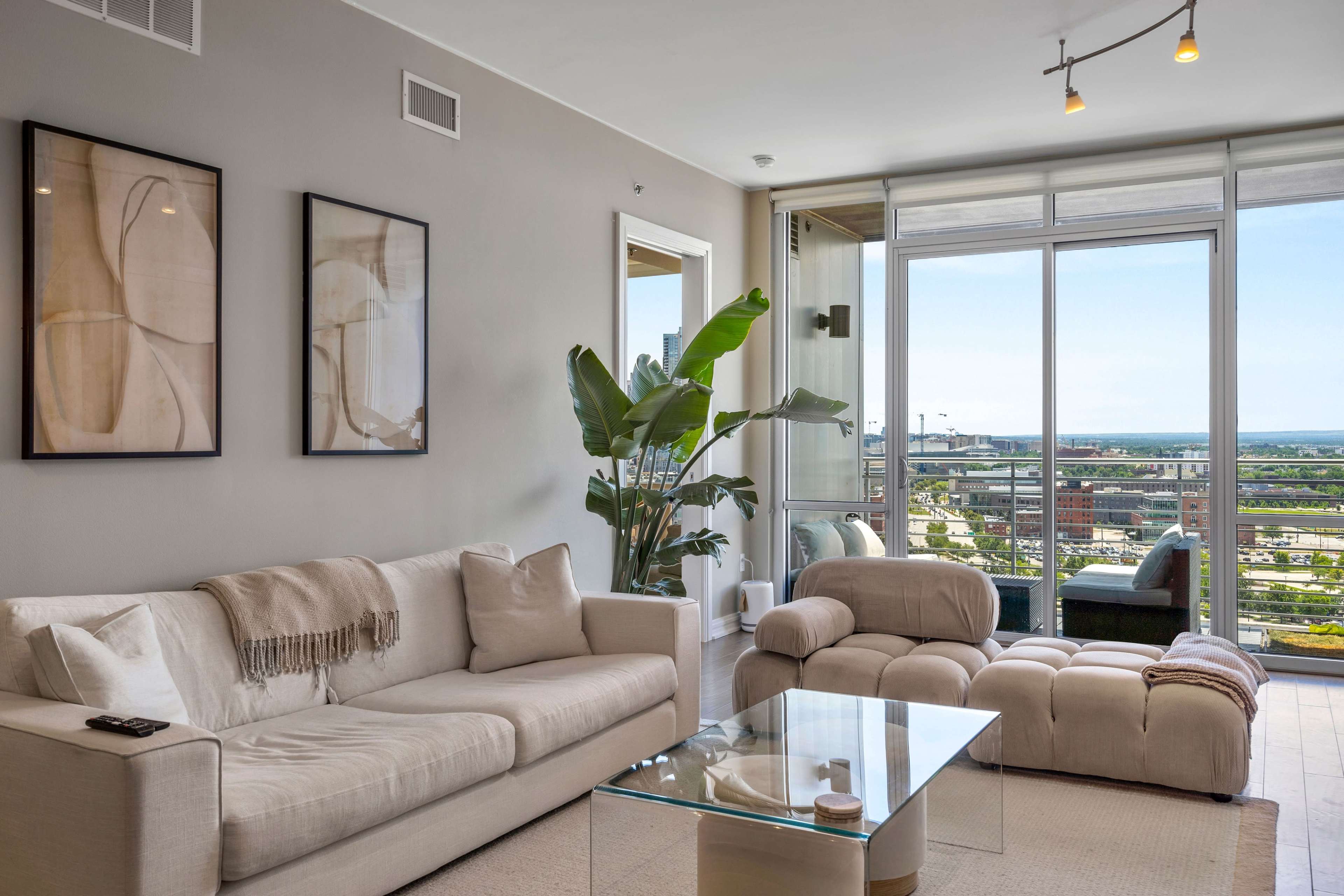 A modern living room features a beige sectional sofa, a glass coffee table, and large windows with a view of the city.