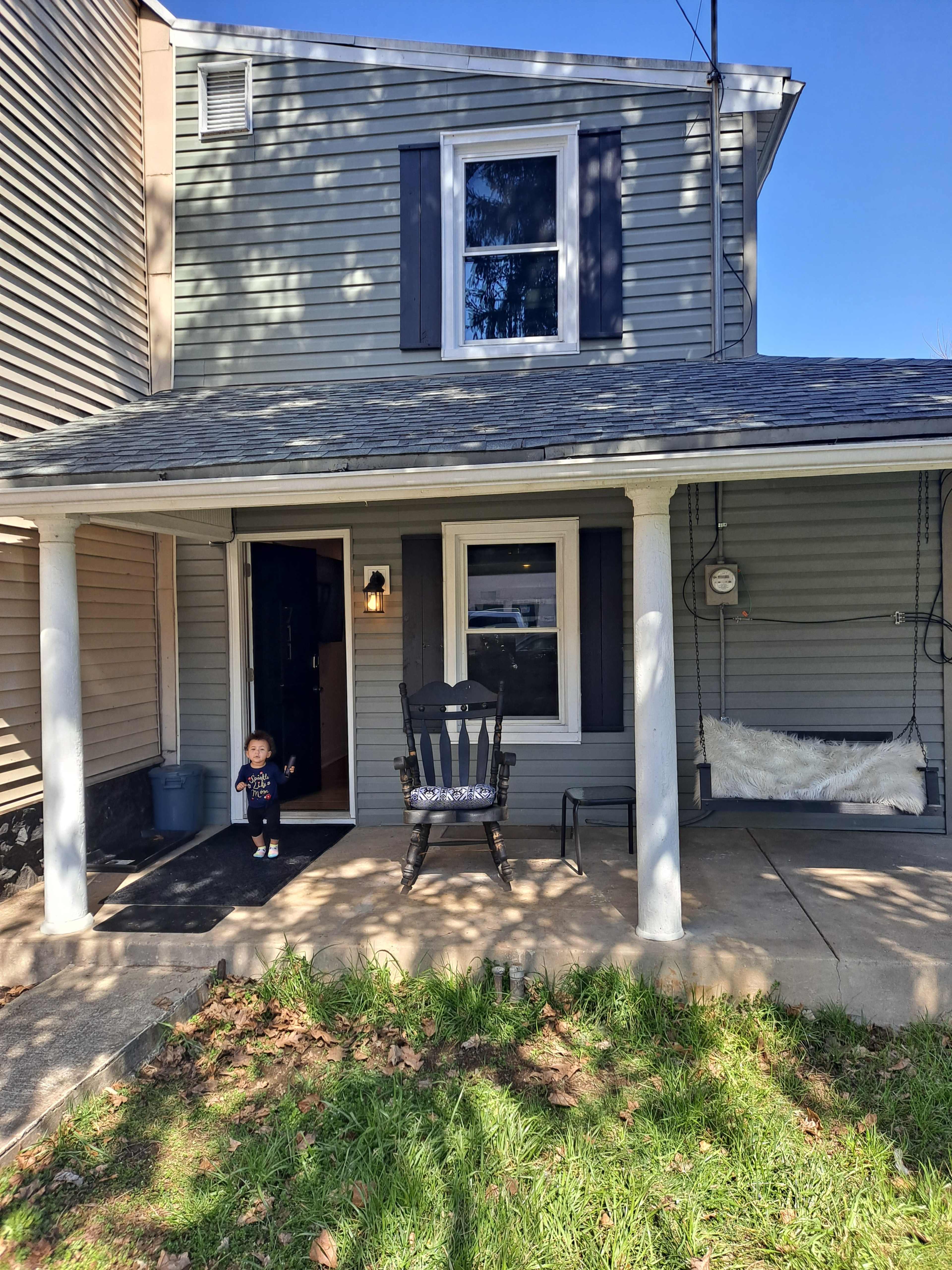 A child stands in front of a two-story house with blue shutters and a porch featuring a rocking chair.