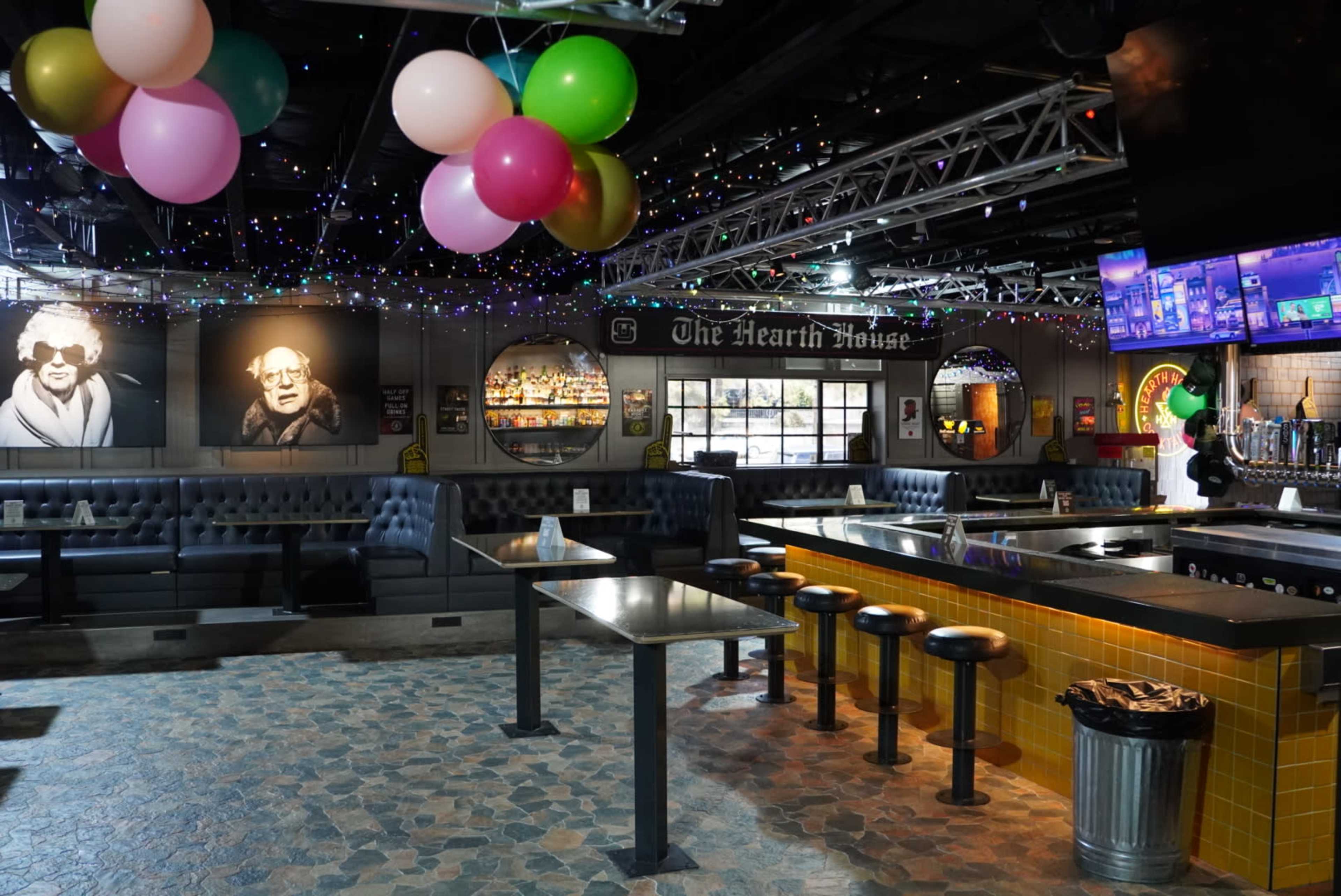 The interior of a bar featuring a central seating area, a long bar counter, and colorful balloons overhead, with dim lighting and decorative photos on the walls.