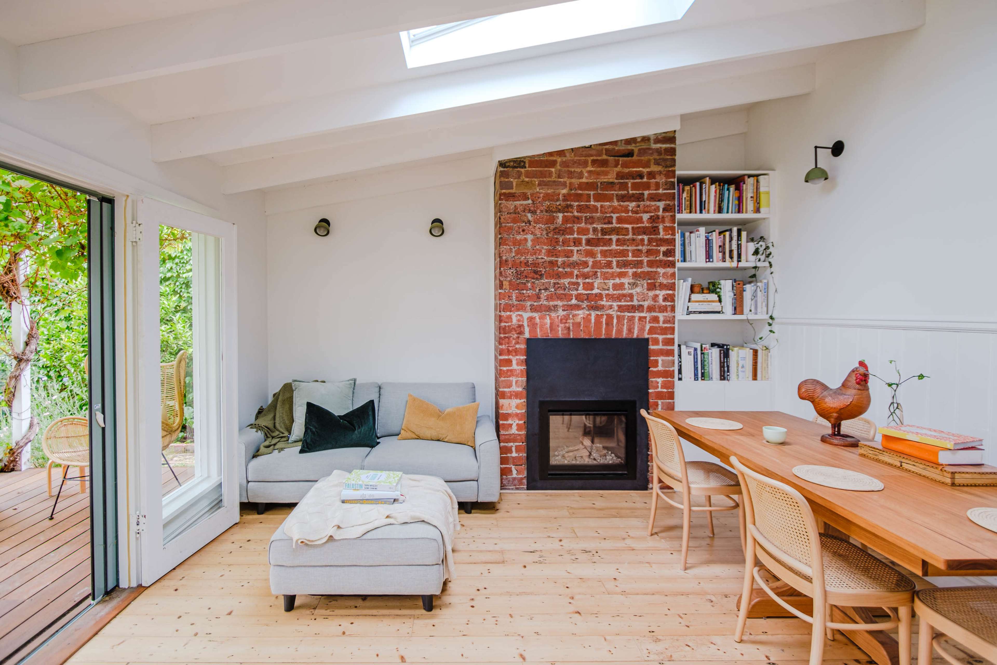 A cozy living room features a gray sofa with a throw pillow, a brick fireplace, a wooden dining table, and a bookshelf against a white wall.