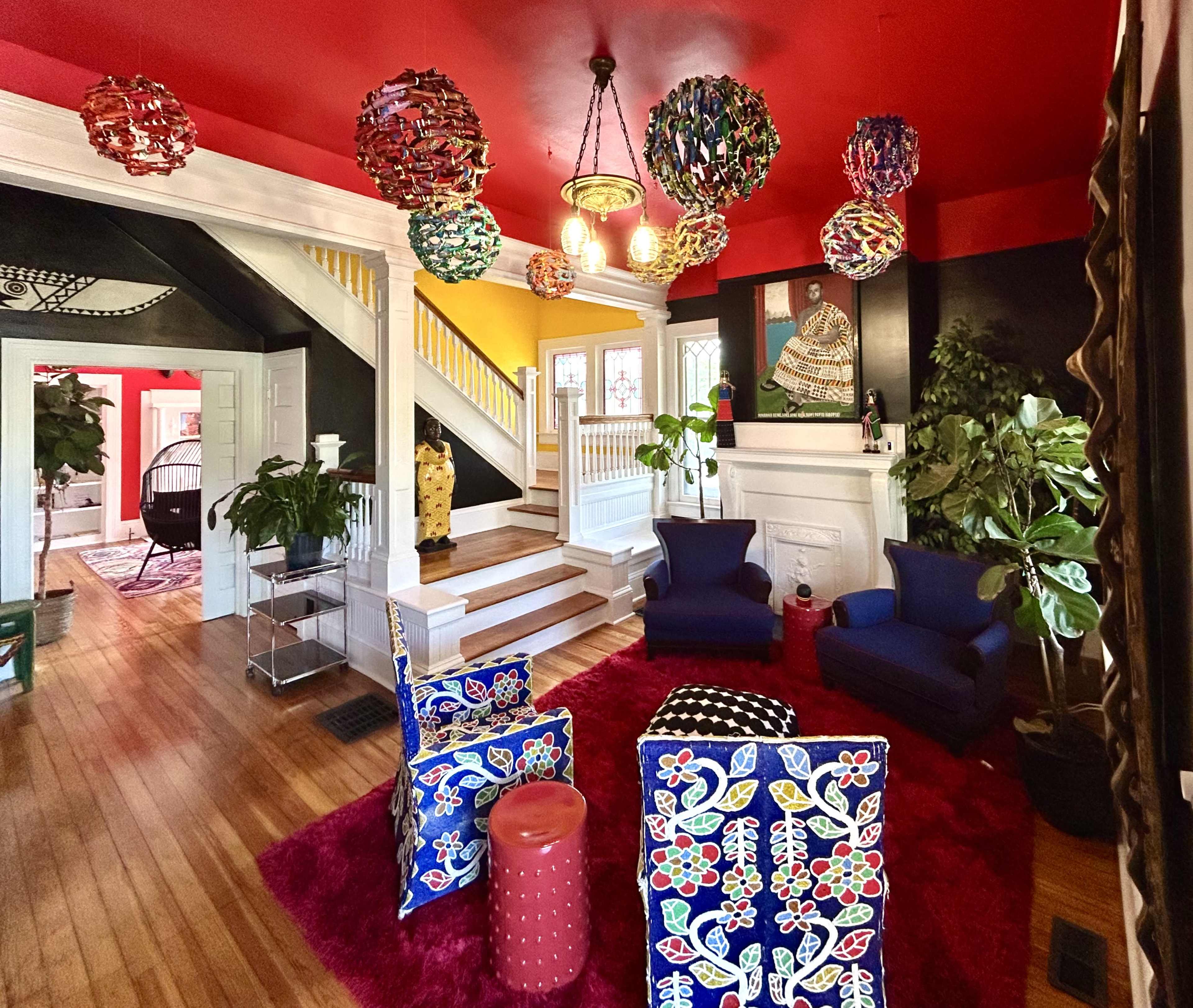 The image shows a vibrant living room with colorful pendant lights, floral-patterned chairs, and a red ceiling, surrounded by potted plants and a staircase.