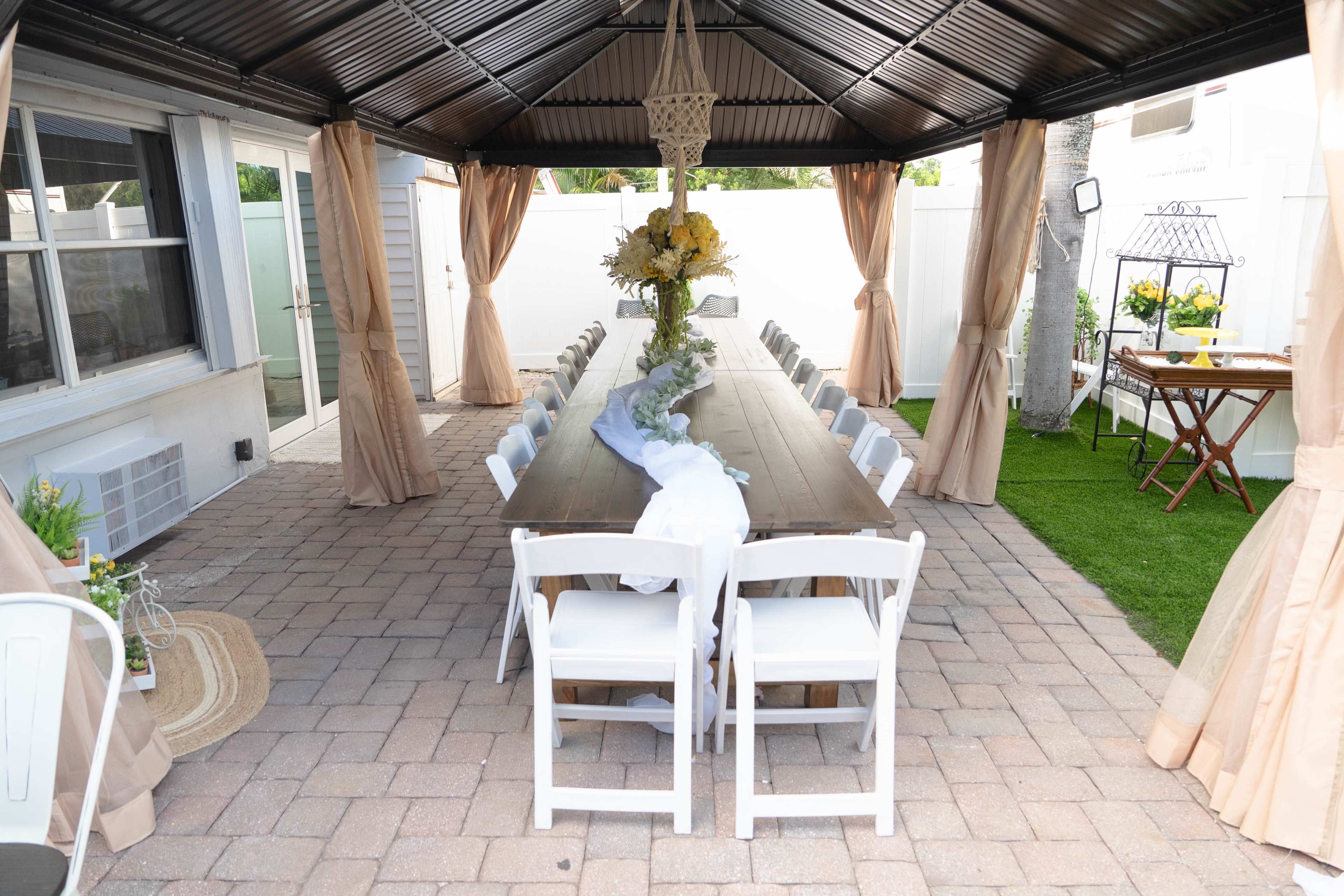 A long wooden table is set under a gazebo adorned with white chairs, while floral arrangements and decorative elements are positioned around the outdoor space.
