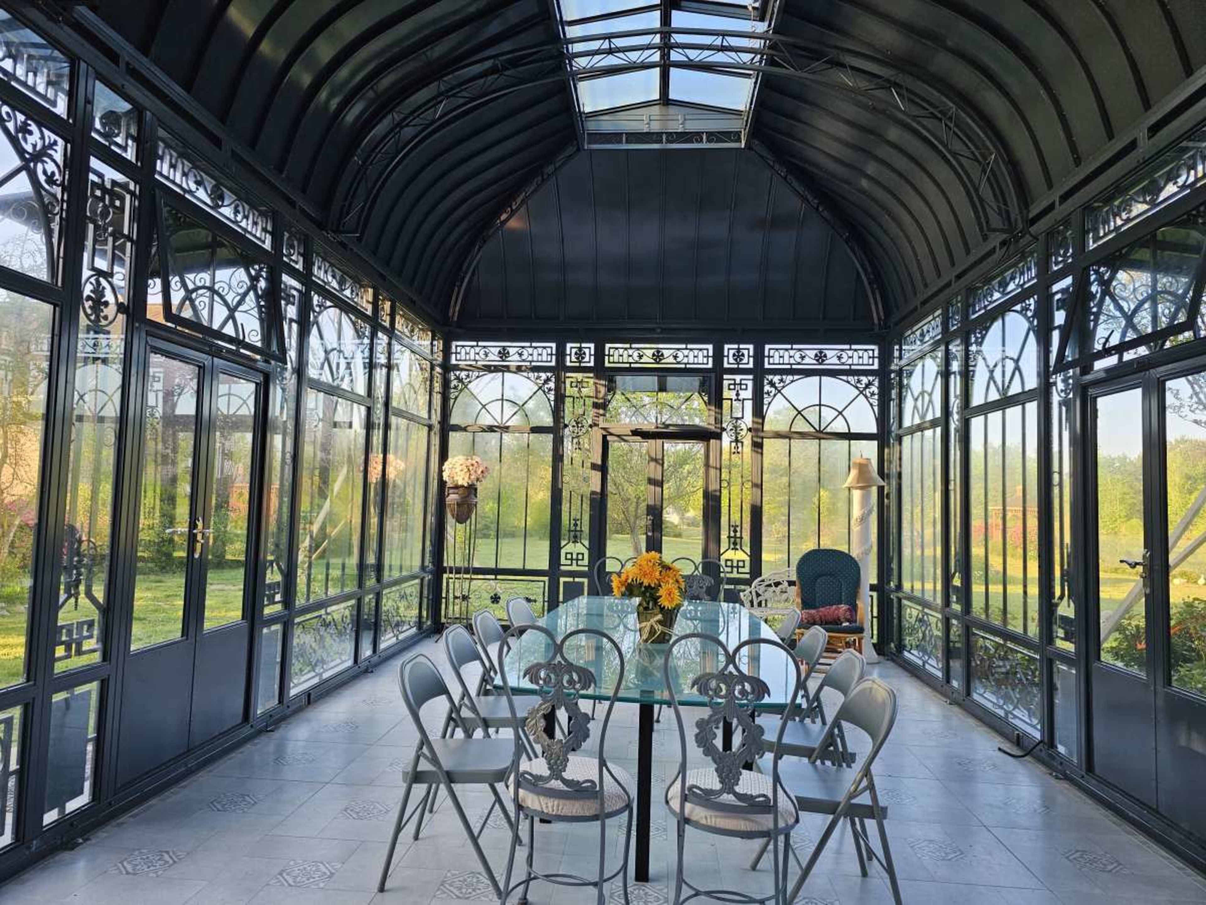 A glass-enclosed conservatory features a dining table surrounded by chairs, with floral arrangements and green views visible through the large windows.