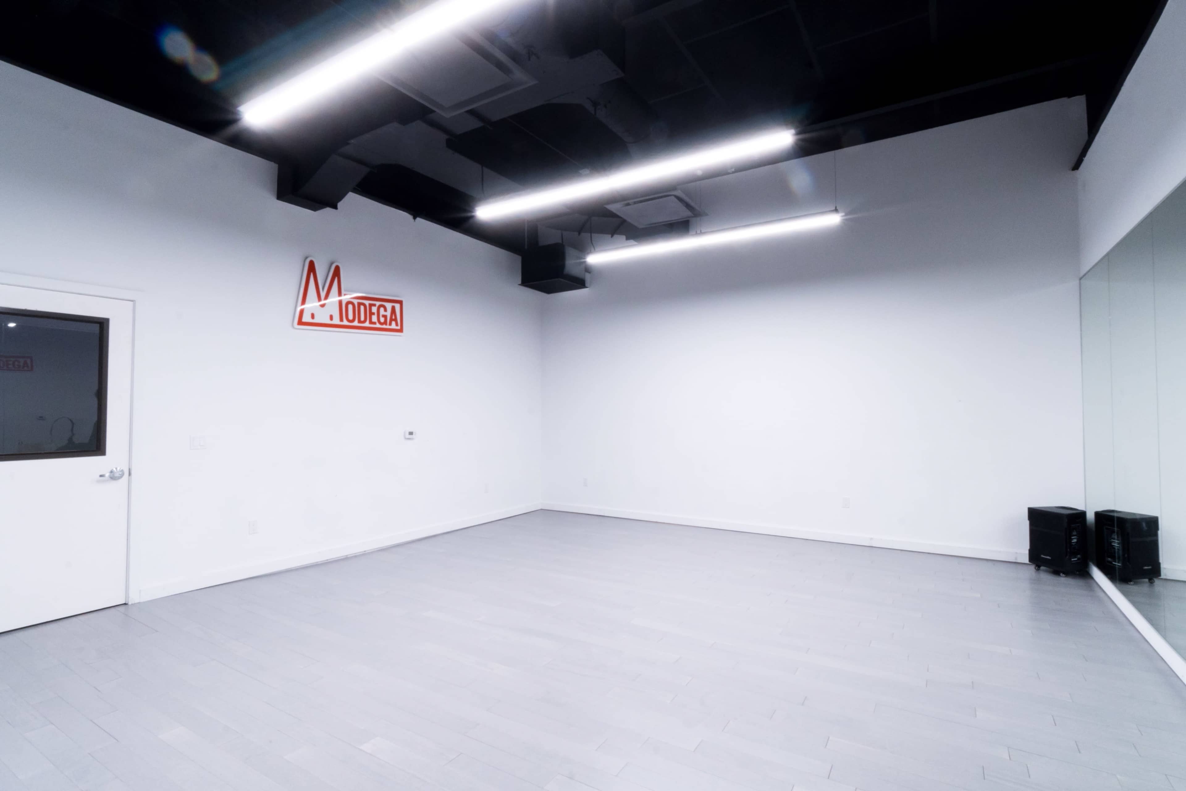 The image shows a nearly empty room with white walls, gray flooring, a door on the left, and a large mirror along the right side, featuring a neon sign that reads "MODEGA."