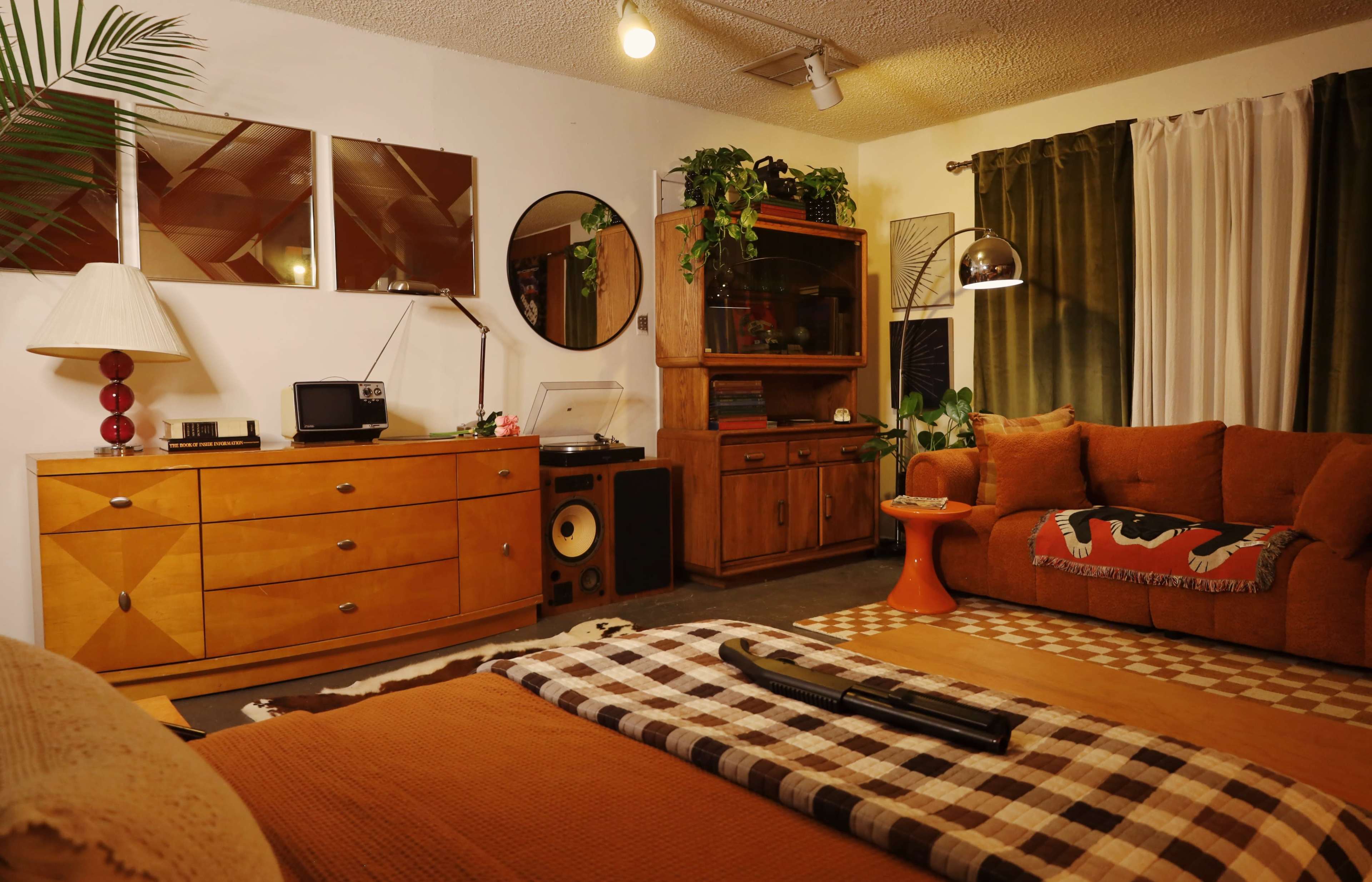 The image depicts a mid-century modern living room featuring a wooden dresser, a record player, a bookshelf, a couch with orange upholstery, and a coffee table with a patterned cloth.