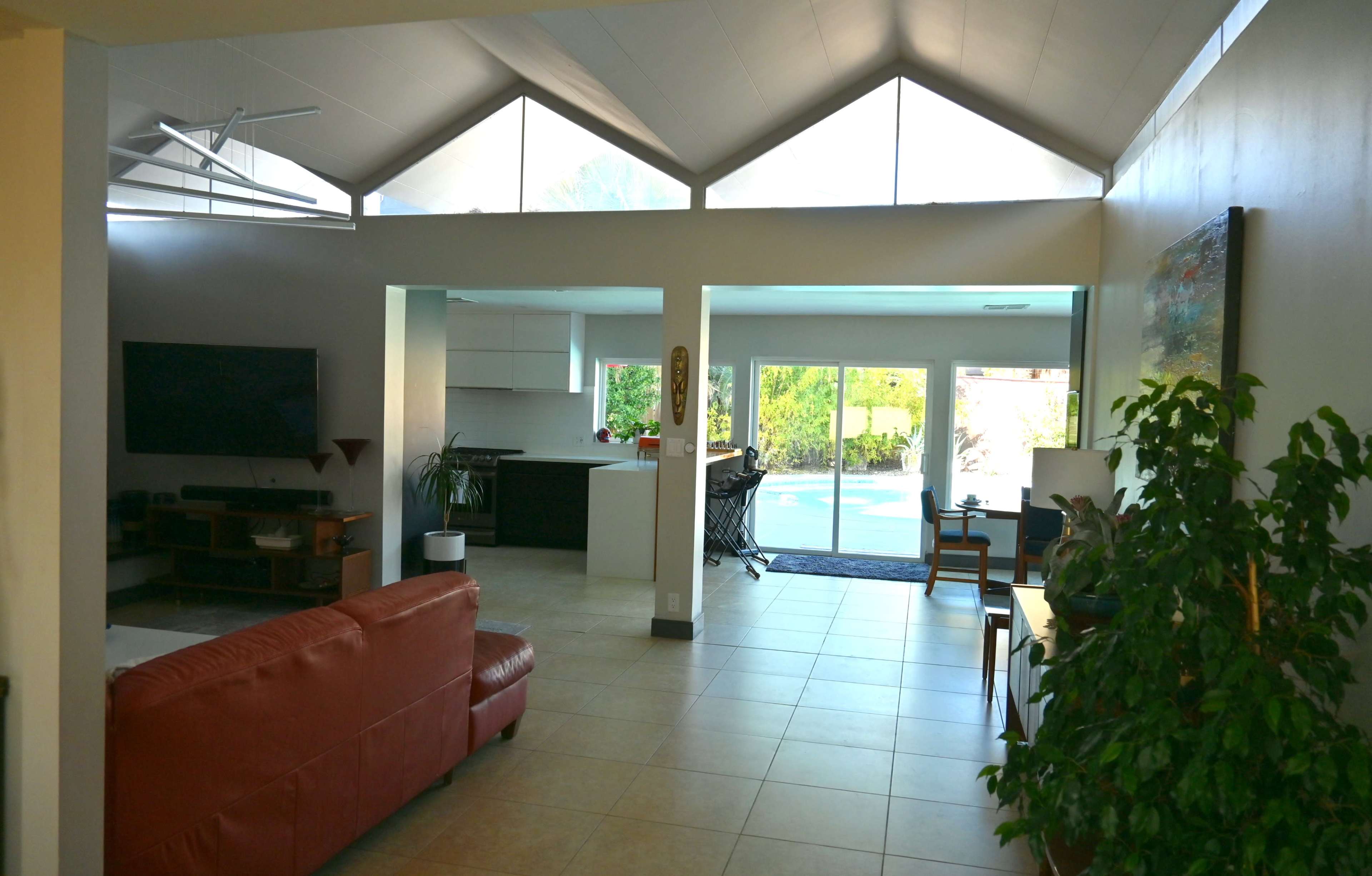 The image shows a modern open-concept living space featuring a living area with a red couch, a kitchen visible in the background, and large windows that open to a poolside view.