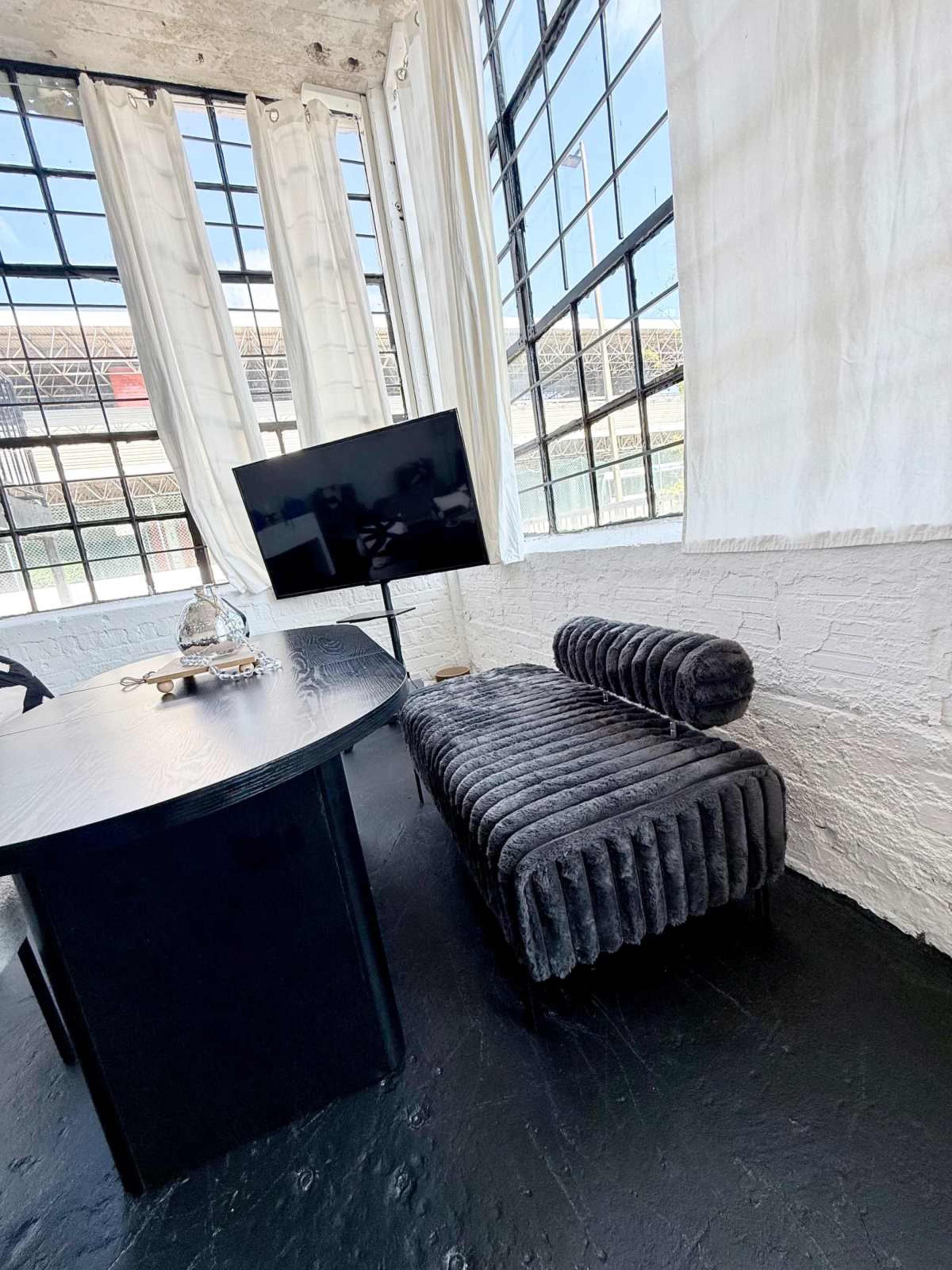 The image shows a modern interior with a dark, plush sofa positioned beside a large window, a black table, and a mounted television.