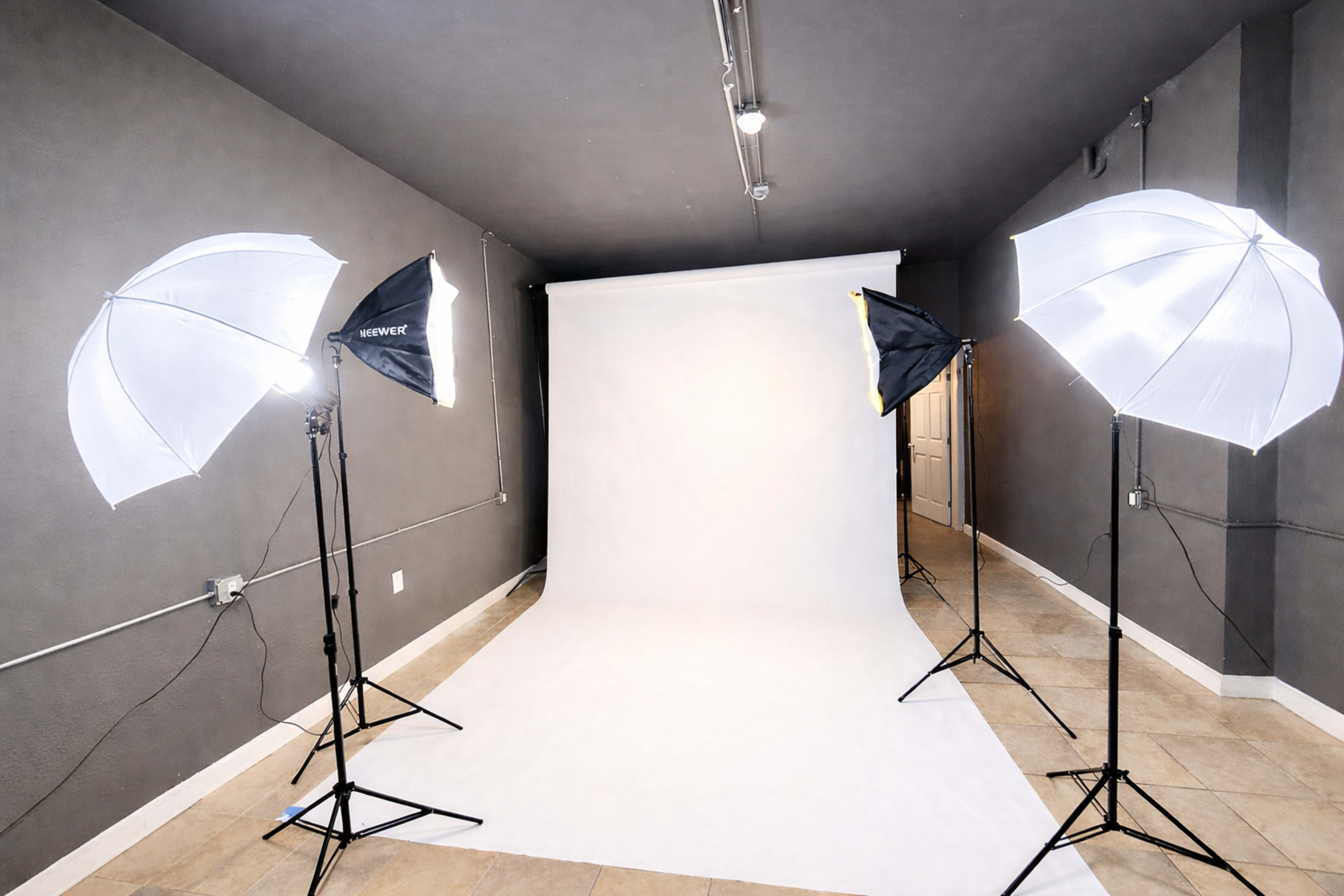 A photography studio features a white backdrop with four light umbrellas positioned around it on a tiled floor.