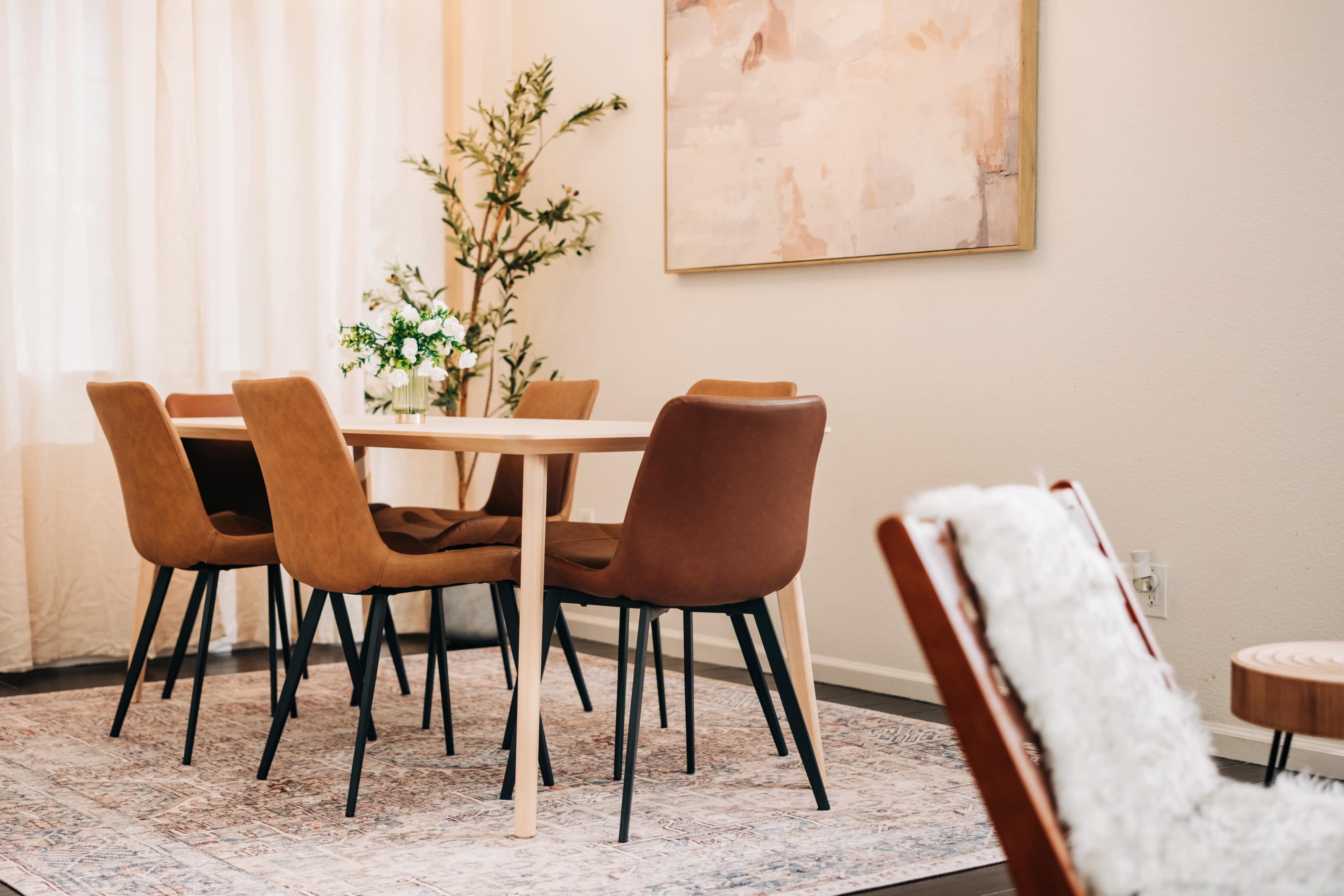 A dining area features a wooden table surrounded by four brown chairs, with a small floral arrangement on the table and a large artwork on the wall.
